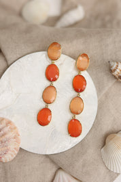 Boucles d'oreilles pendantes dorées Adrienne orange ornées de pierres œil de chat - Bijou photographié dans un décor estival