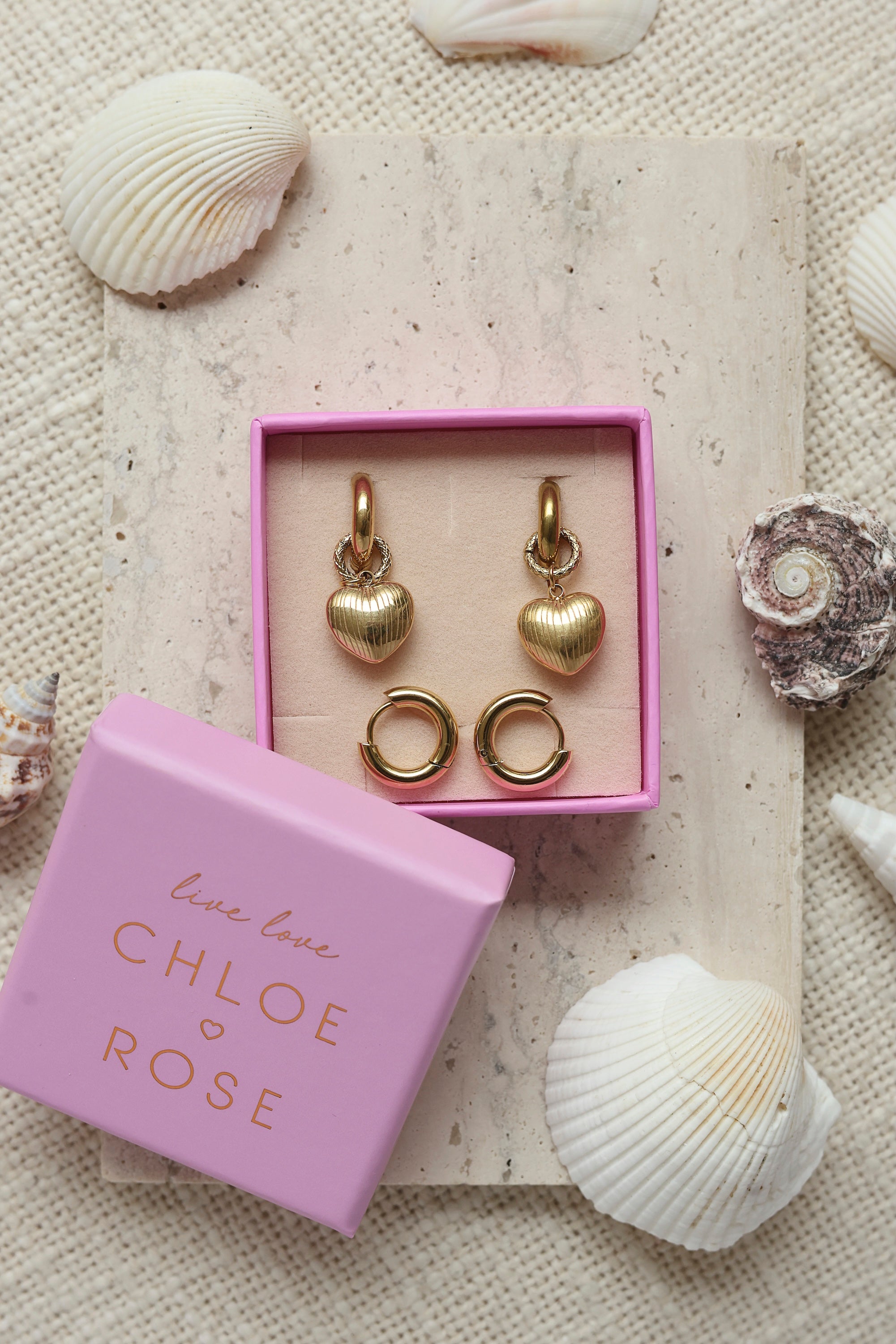 Coffret de parure de bijoux Balmy comprenant une paire de mini créoles dorées ornées d’un cœur strié ainsi qu’une paire de créoles simples - Bijoux photographiés dans un décor estival