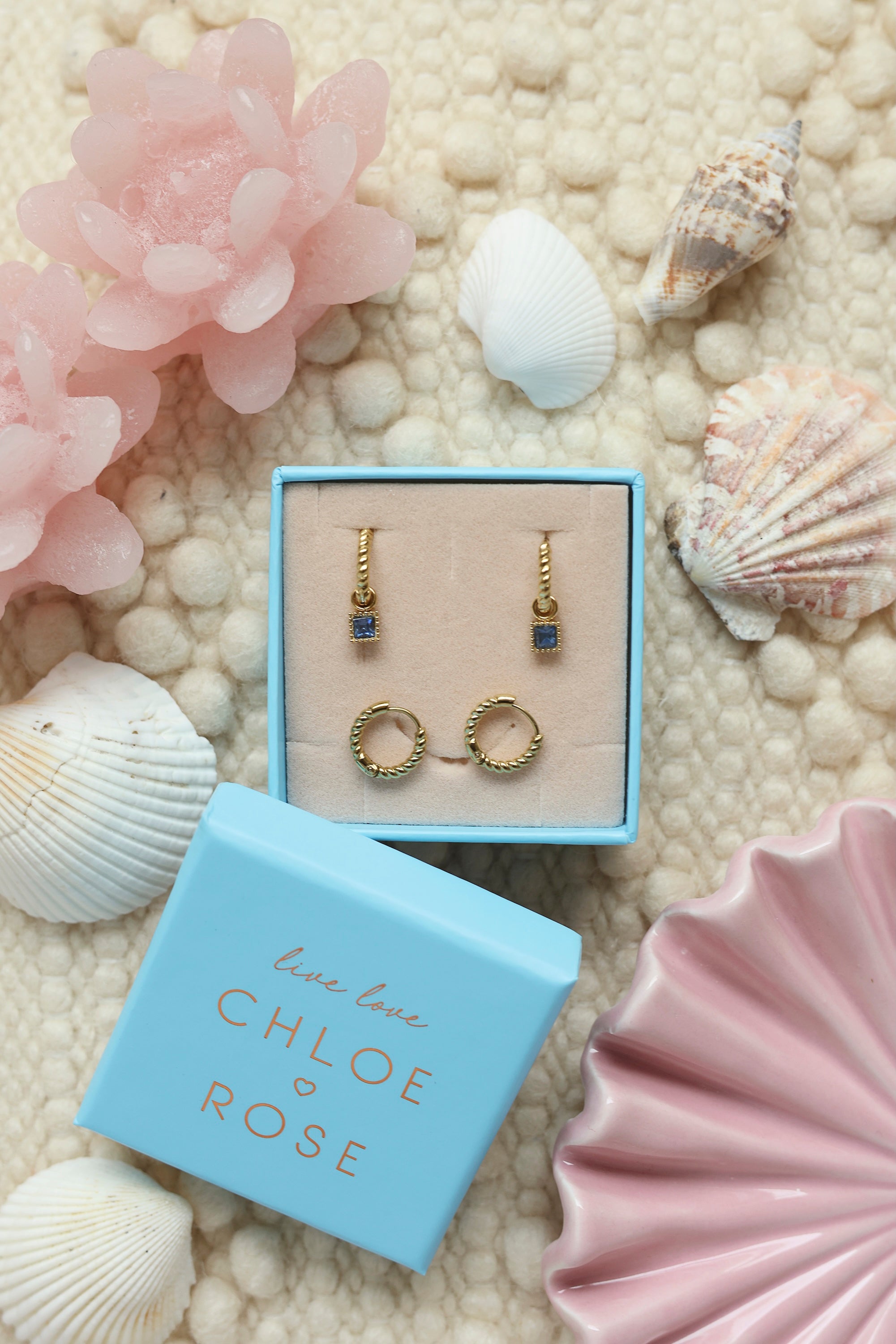 Coffret de parure de bijoux Lush bleu comprenant une paire de mini créoles torsadées dorées ornées d’un zircon carré bleu ainsi qu’une paire de créoles torsadées simples - Bijoux photographiés dans un décor estival