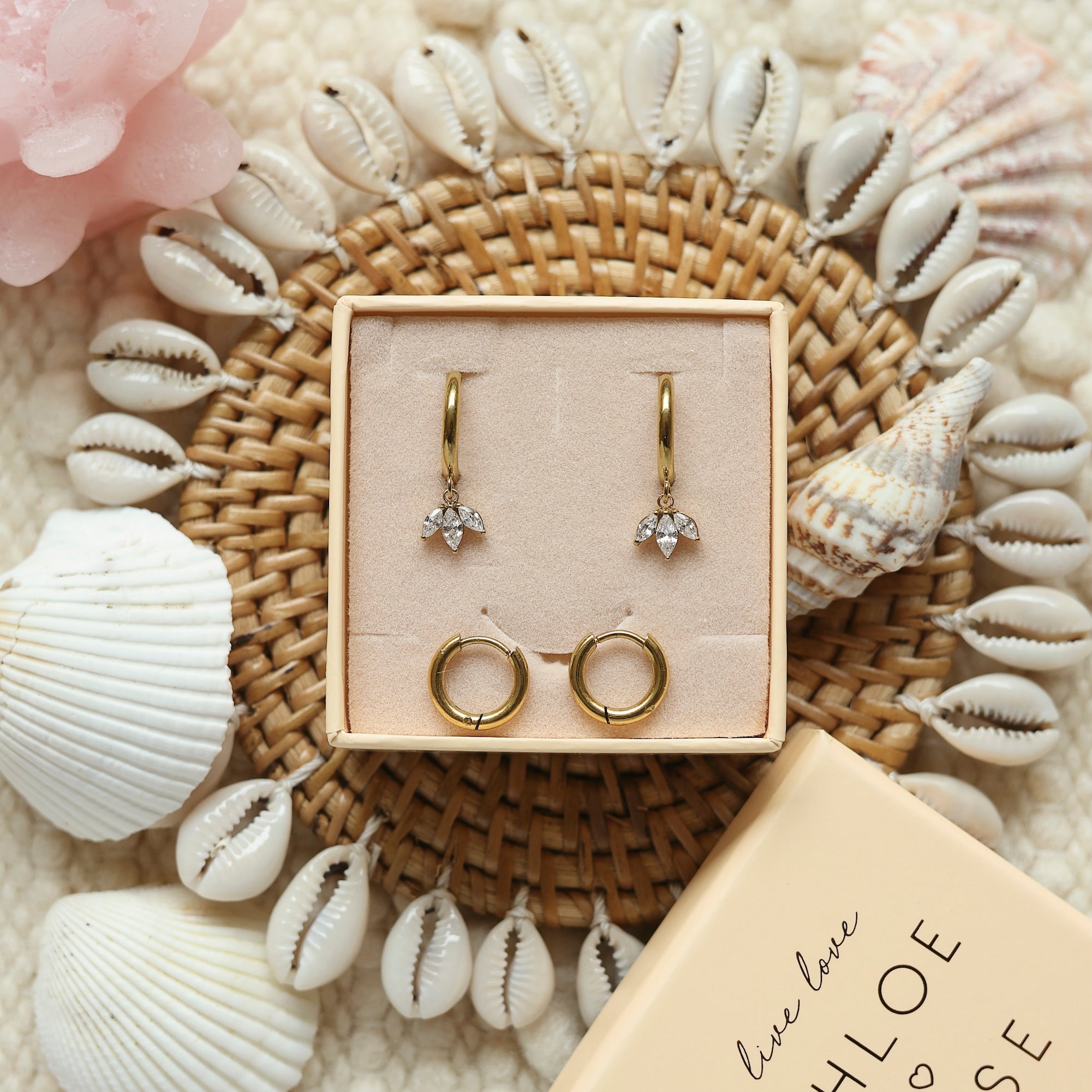 Coffret de parure de bijoux Mirage comprenant une paire de mini créoles dorées ornées d’un pendentif pétales serti de zircons ainsi qu’une paire de créoles simples - Bijoux photographiés dans un décor estival
