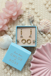 Coffret de parure de bijoux Oasis bleu comprenant une paire de mini créoles dorées ornées d’un pendentif pétales serti de zircons ainsi qu’une paire de créoles simples et une paire de puces assorties - Bijoux photographiés dans un décor estival