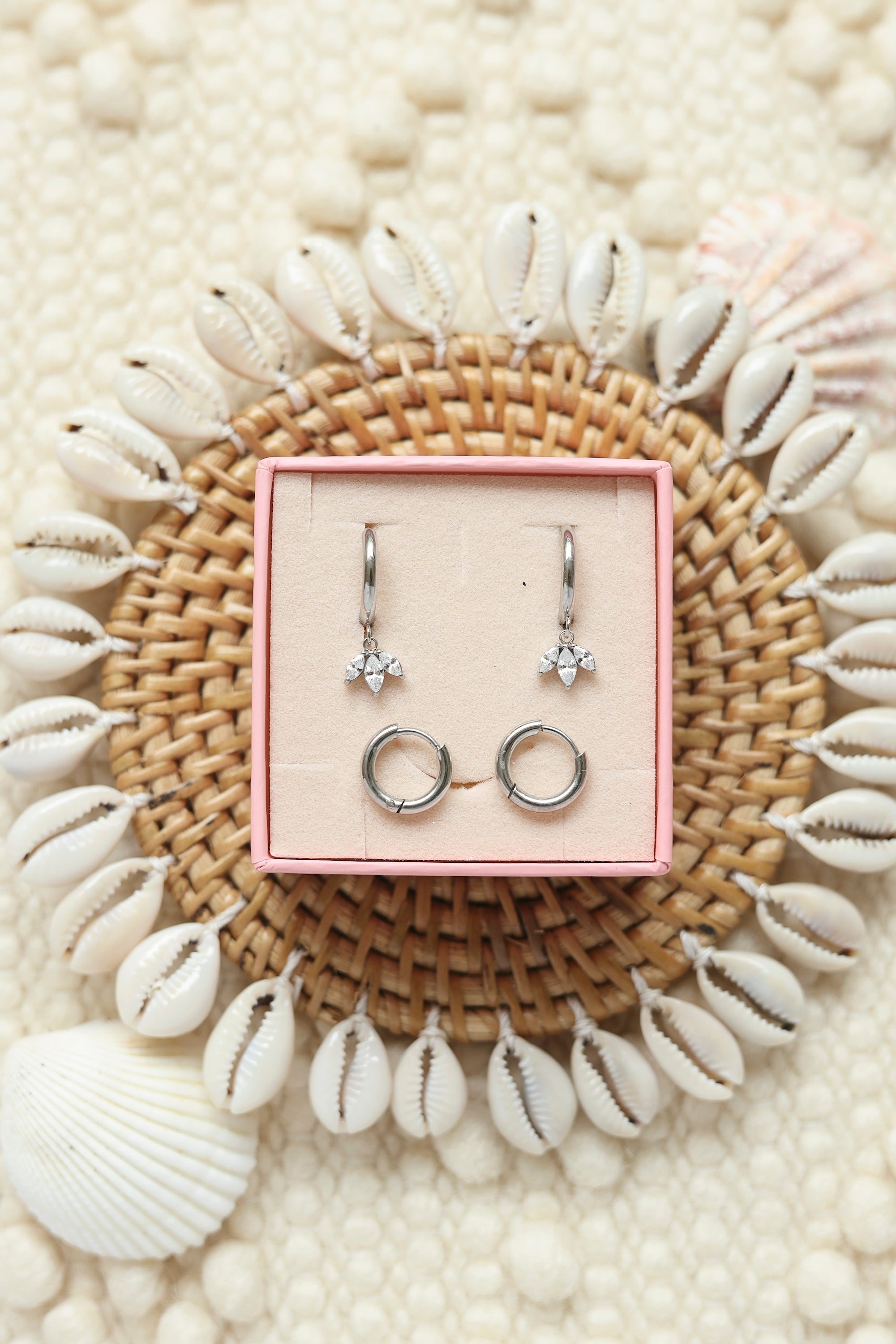 Coffret de parure de bijoux Mirage comprenant une paire de mini créoles argentées ornées d’un pendentif pétales serti de zircons ainsi qu’une paire de créoles simples - Bijoux photographiés dans un décor estival
