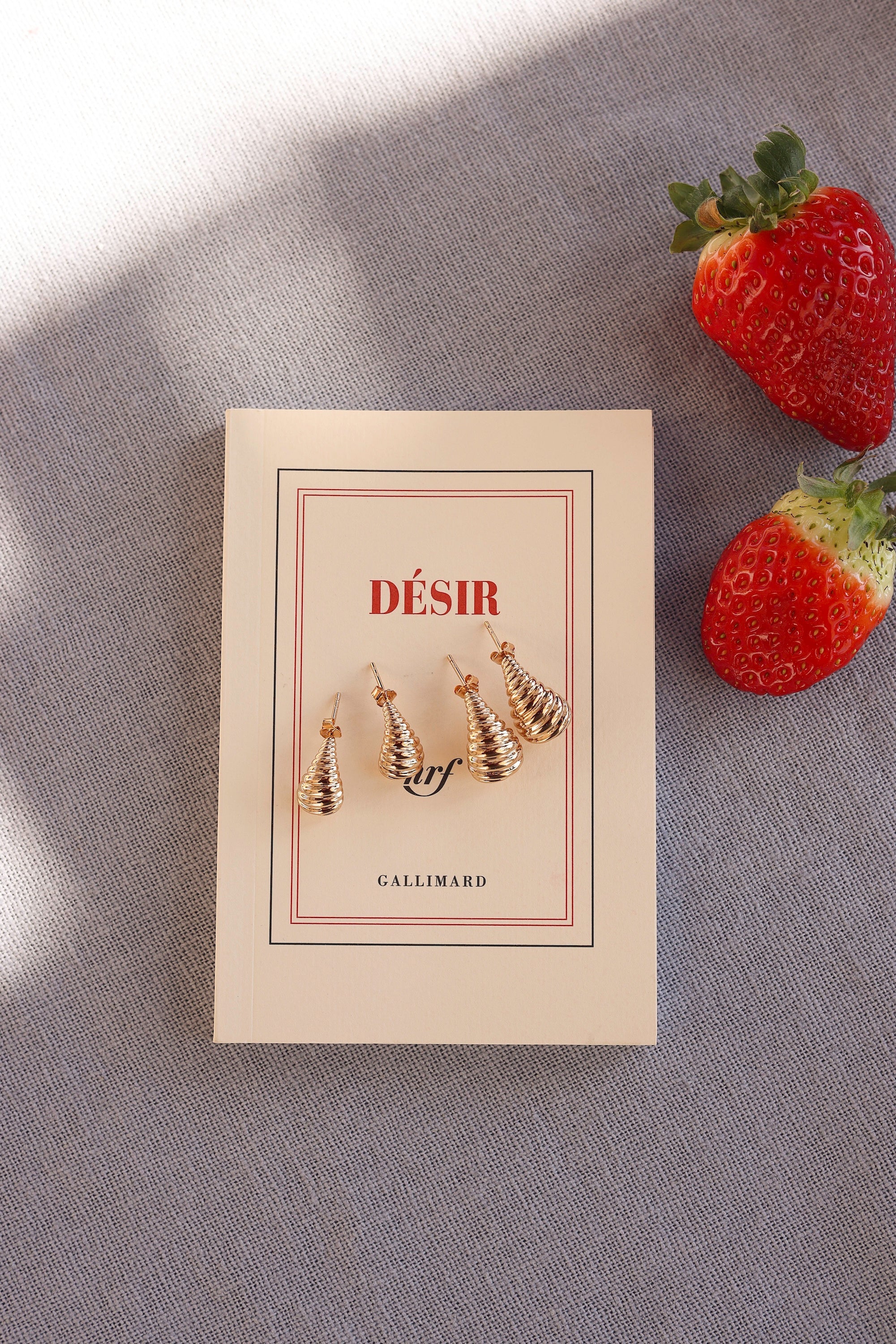 Boucles d'oreilles dorées Clelia S - Bijou photographié sur livre et tissu bleu accompagné de fraise