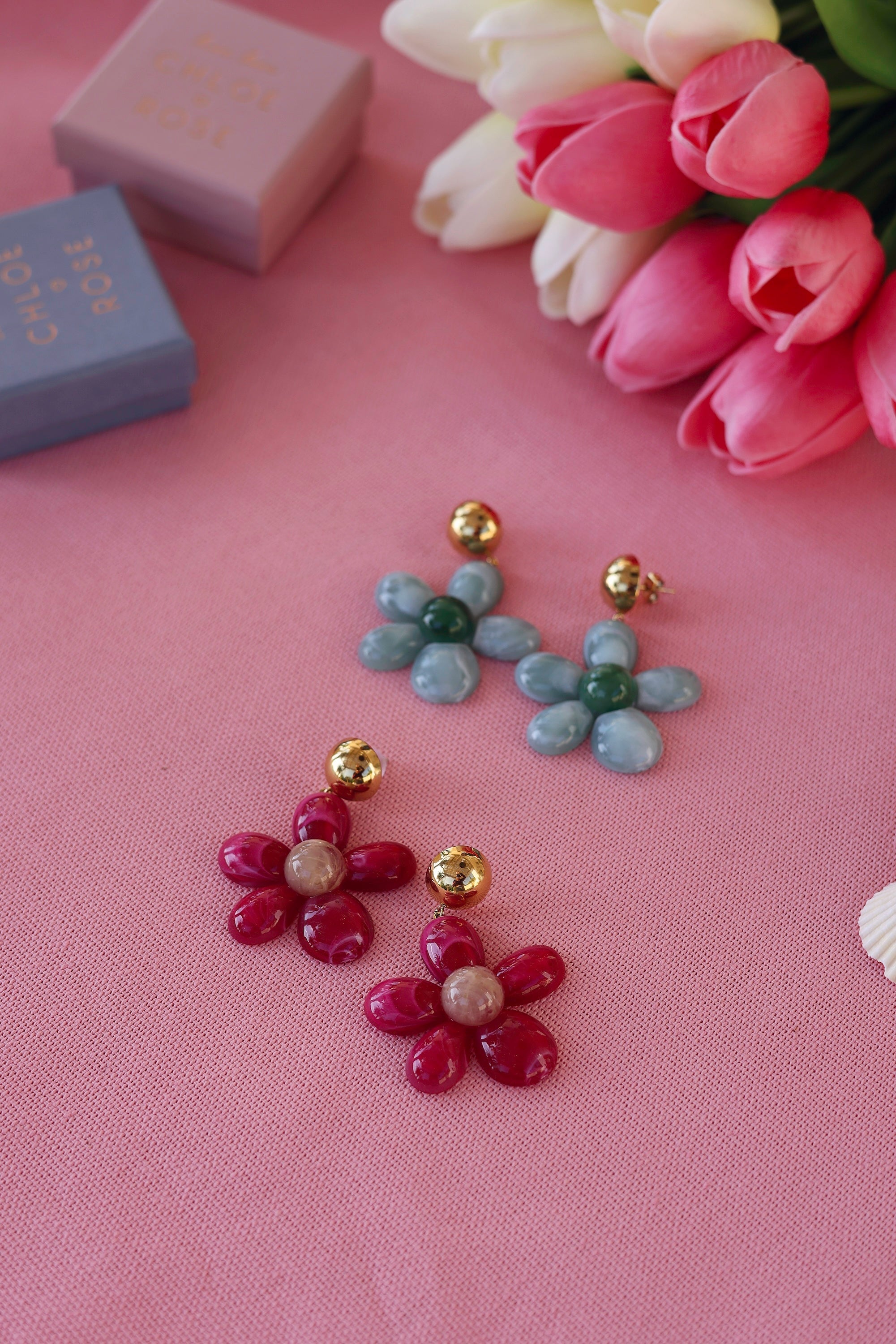 Boucles d'oreilles pendantes dorées Marty fuchsia en acétate - Bijou photographié sur tissu rose avec tulipes et boites cadeau Chloe Rose