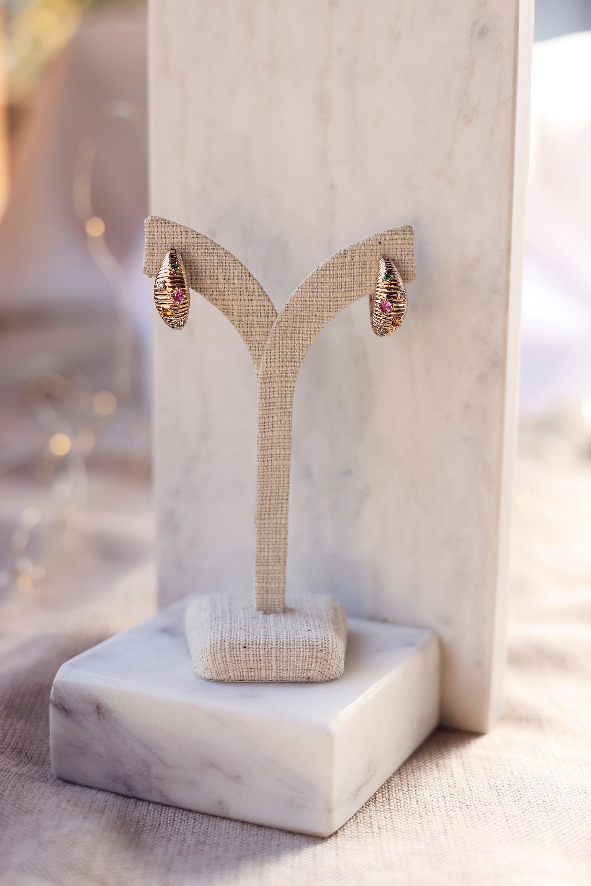 Boucles d'oreilles créoles dorées Janina striées et ornées de zircons colorés - Bijou photographié sur un présentoir en lin