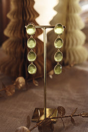 Boucles d'oreilles pendantes dorées Adrienne ornées de pierres naturelles - Bijou photographié sur un présentoir doré avec en fond des décorations de Noël