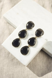 Boucles d'oreilles pendantes dorées Gisèle ornées de cristaux effet oeil de chat - Bijou photographié sur un présentoir en marbre blanc posé sur un tissu en lin beige