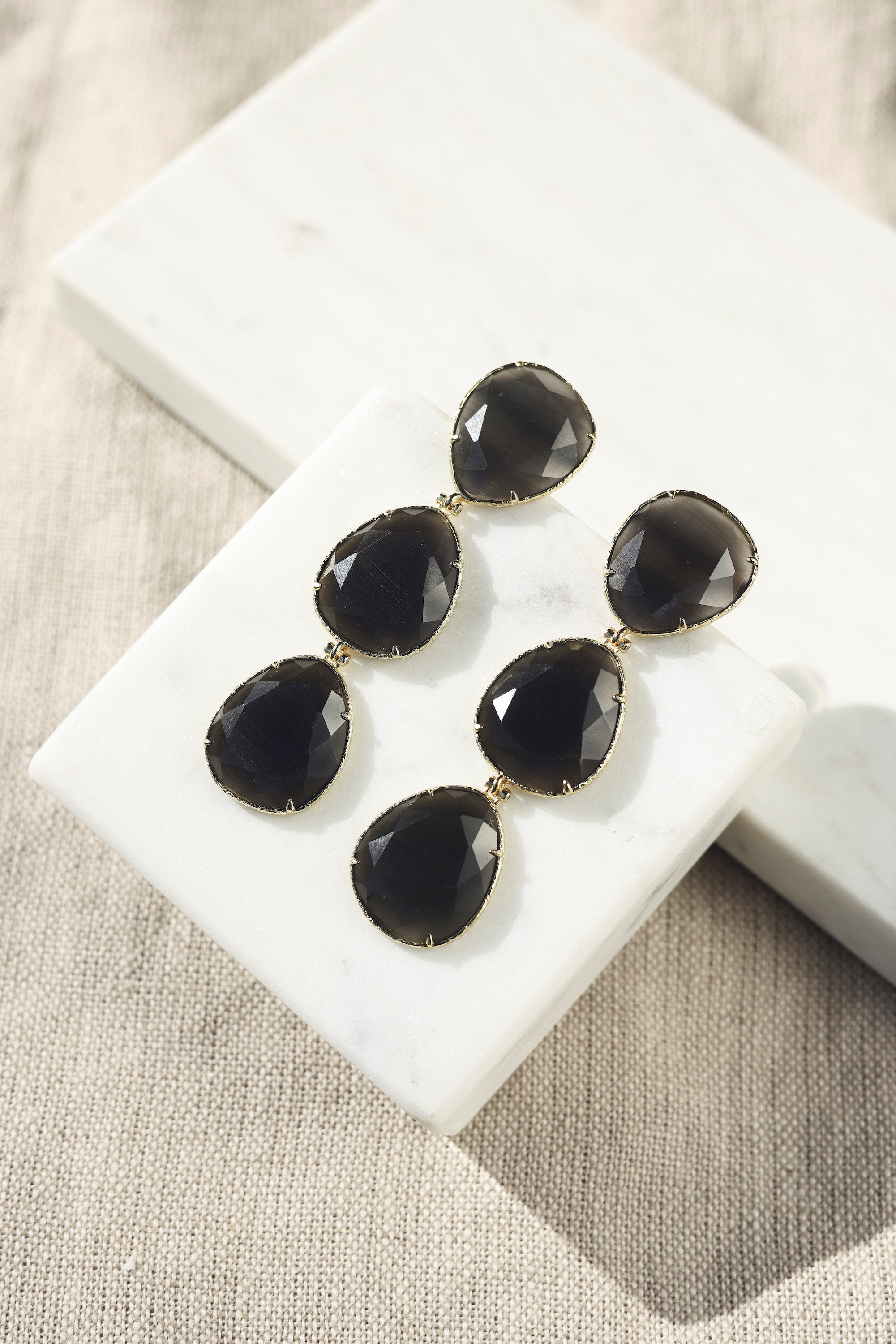 Boucles d'oreilles pendantes dorées Gisèle ornées de cristaux effet oeil de chat - Bijou photographié sur un présentoir en marbre blanc posé sur un tissu en lin beige