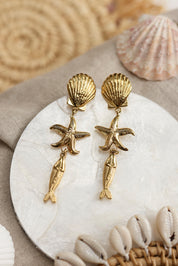 Boucles d'oreilles pendantes dorées Keliane coquillage, étoile de mer et poisson - Bijou photographié dans un décor estival