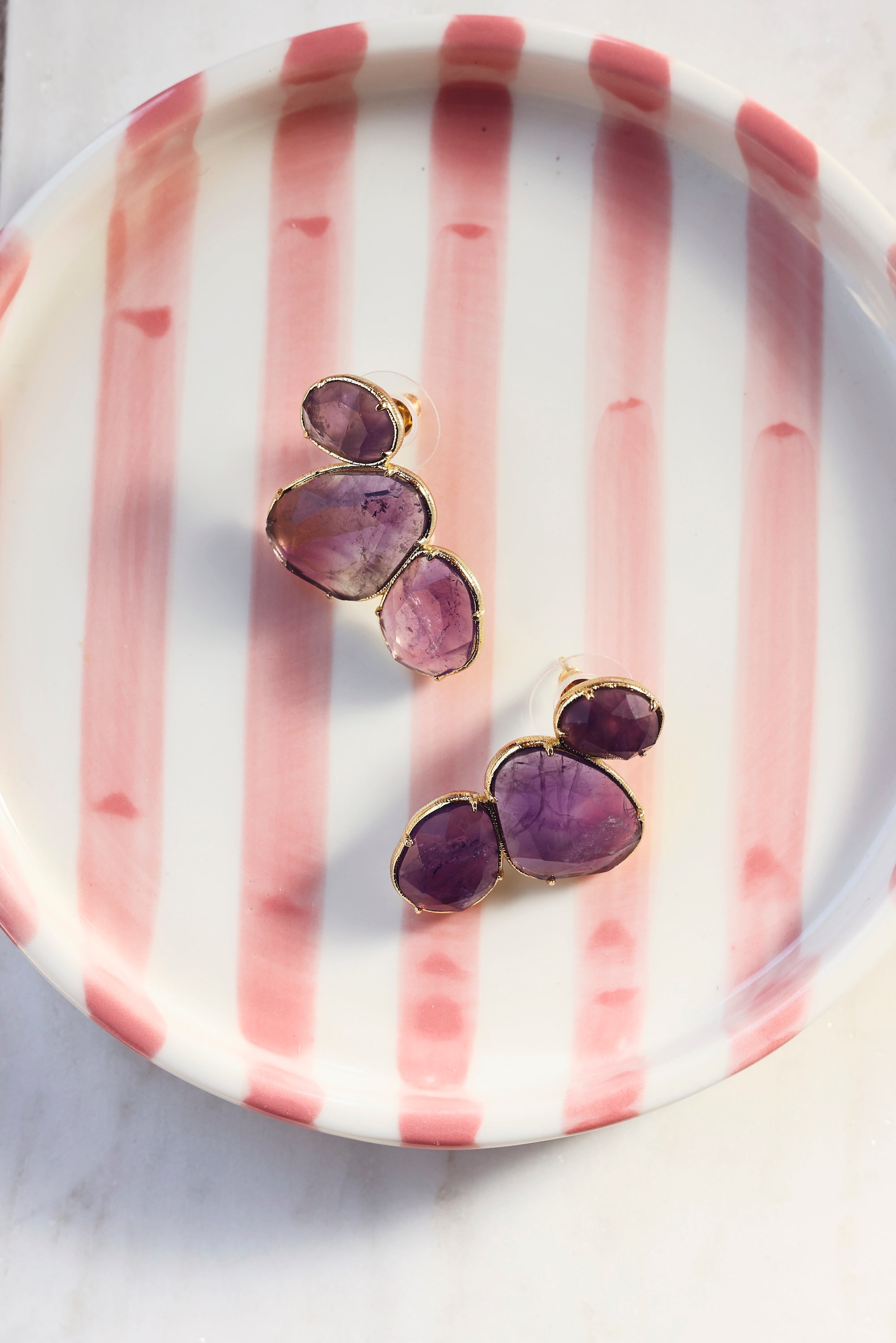 Boucles d'oreilles clous Lindsay améthyste dorées ornées de pierres naturelles - Bijou photographié dans un vide poche rose et blanc
