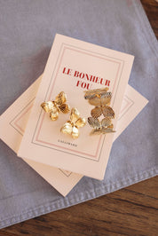 Boucles d'oreilles dorées Jenny papillon - Bijou photographié sur livre "Le bonheur fou" posé sur un tissu en lin bleu sur une table en bois