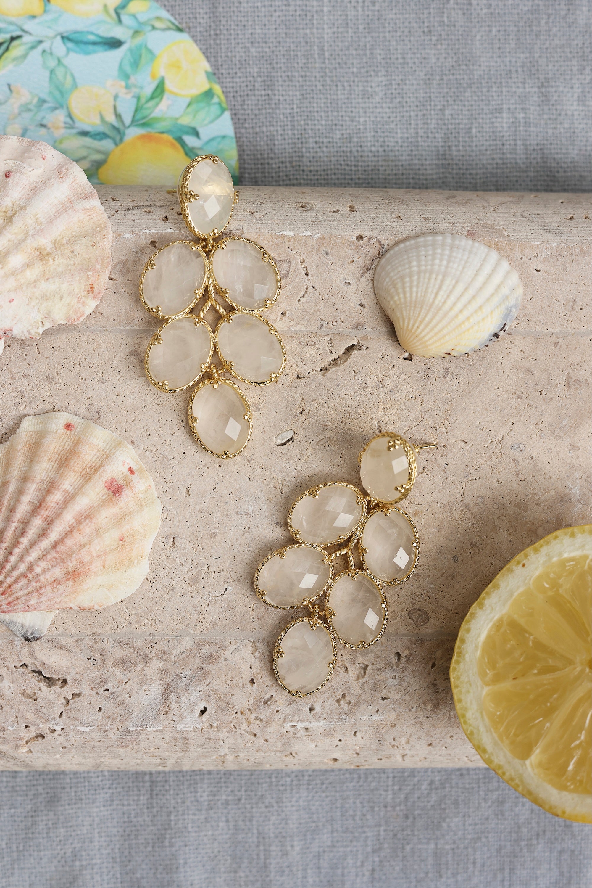 Boucles d'oreilles pendantes dorées Layana motif grappe de raisins ornées de pierres naturelles - Bijou photographié dans un décor estival
