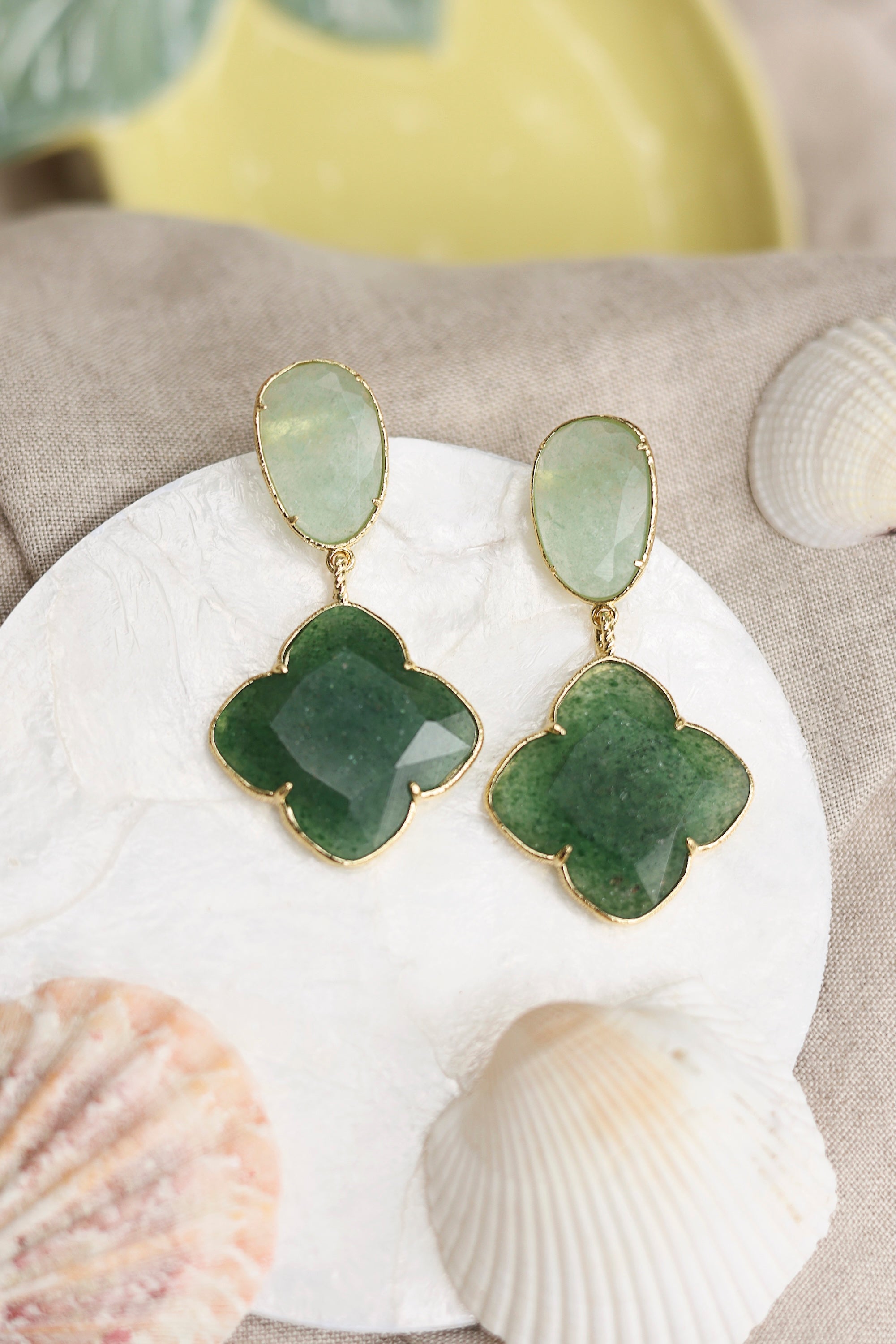 Boucles d'oreilles pendantes dorées Zephira vert à motif trèfle ornées de pierres naturelles -Bijou photographié dans un décor estival