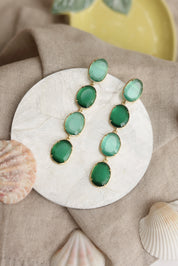 Boucles d'oreilles pendantes dorées Adrienne vert haut ornées de pierres œil de chat - Bijou photographié dans un décor estival