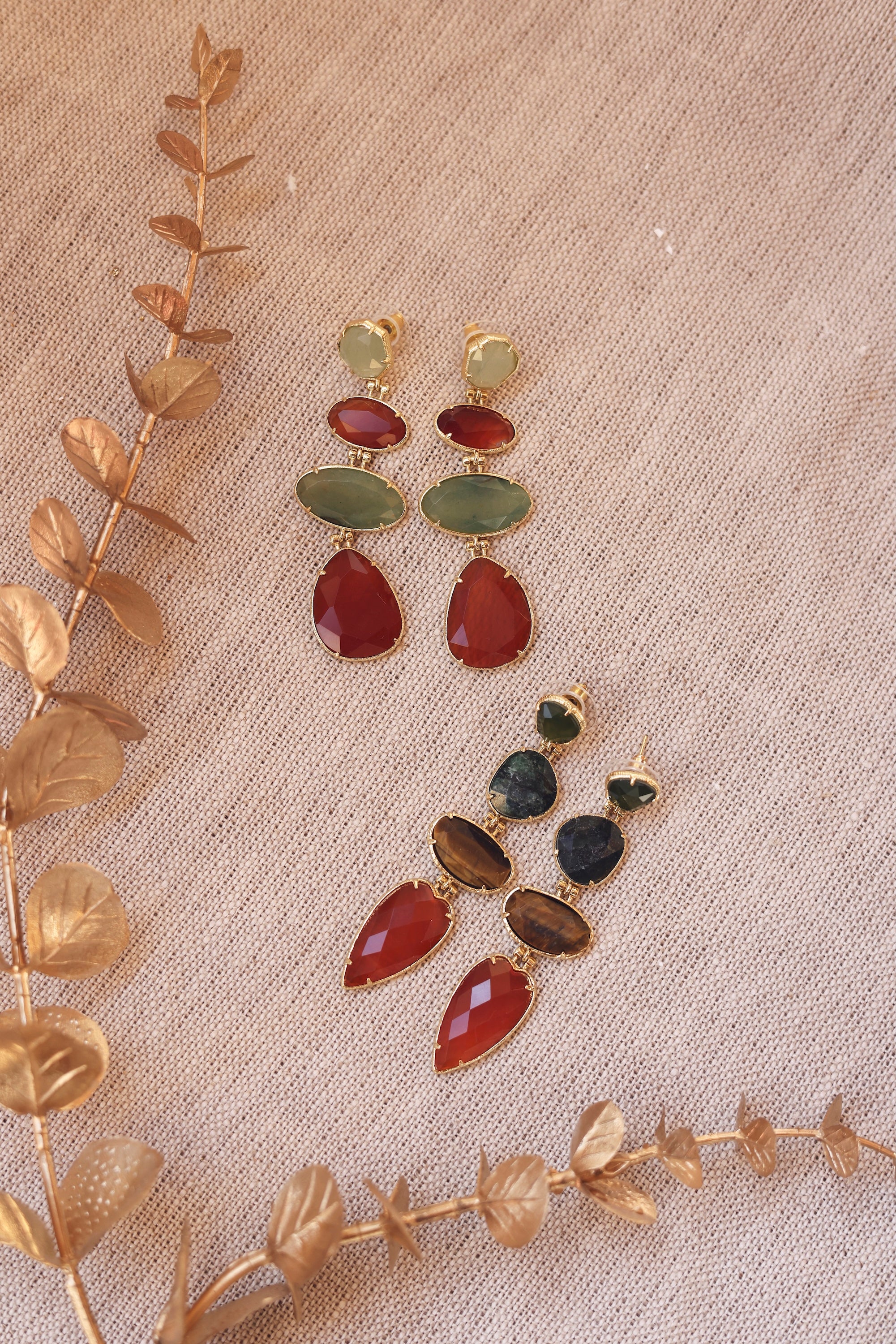 Boucles d'oreilles pendantes dorées Delfina ornées de pierres naturelles - Bijou  photographié sur un tissu en lin beige avec des feuilles dorées