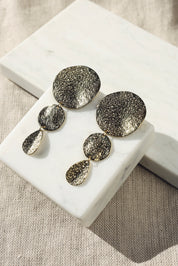 Boucles d'oreilles pendantes dorées Elly martelées - Bijou photographié sur un présentoir en marbre blanc posé sur un tissu en lin beige