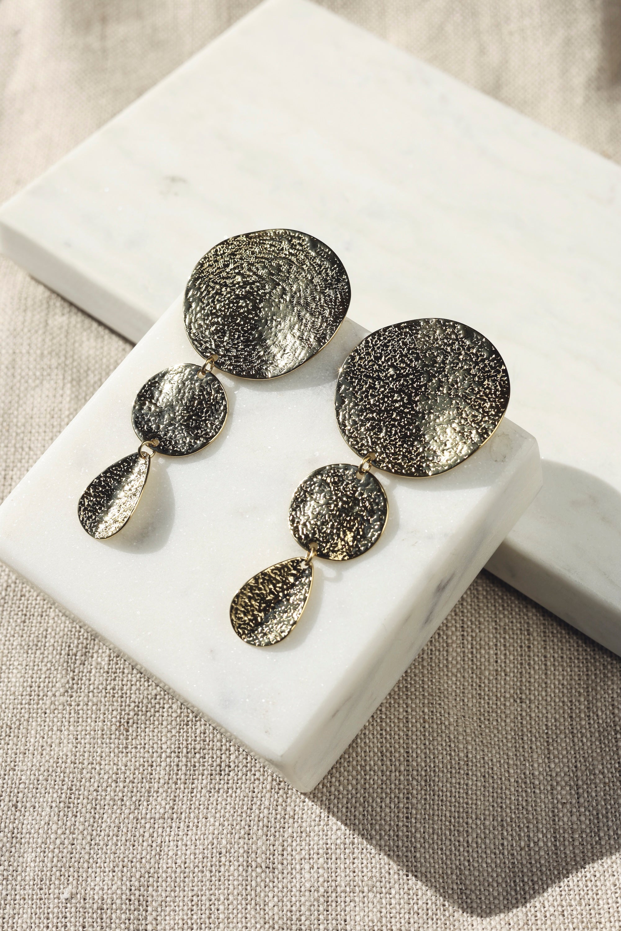 Boucles d'oreilles pendantes dorées Elly martelées - Bijou photographié sur un présentoir en marbre blanc posé sur un tissu en lin beige