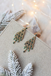 Boucles d'oreilles pendantes dorées Merilia ornées de pierres naturelles disposées en forme de plumes de paon - Bijou photographié sur un présentoir en lin gris dans une ambiance hivernale