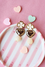Boucles d'oreilles pendantes Zohra beige dorées trèfle, cœur martelé et acétate - Bijou photographié dans une coupole ronde rayée rose et blanche posée sur un tissu rose