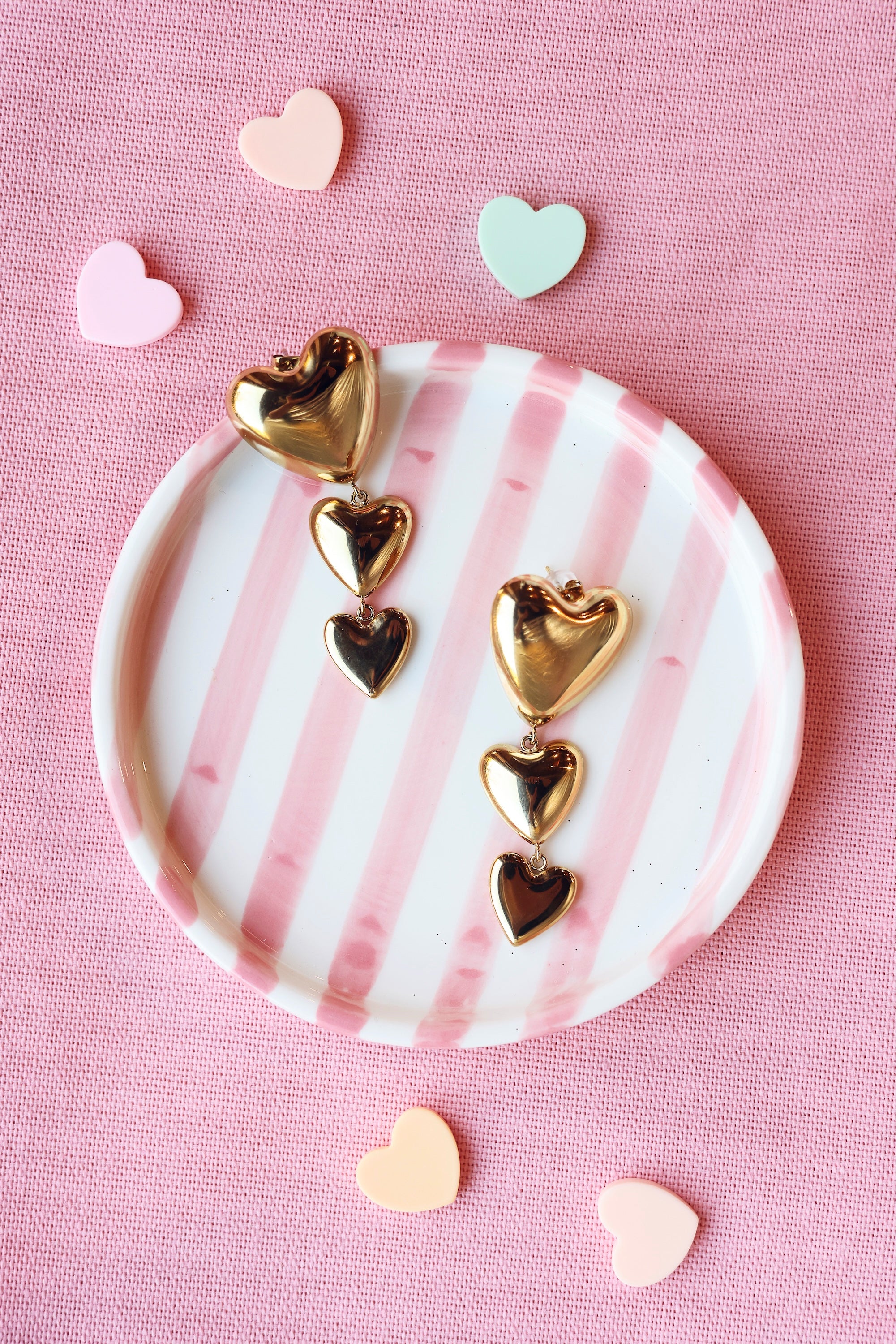 Boucles d'oreilles pendantes Raelle dorées cœurs - Bijou photographié dans une coupole ronde rayée rose et blanche posée sur un tissu rose