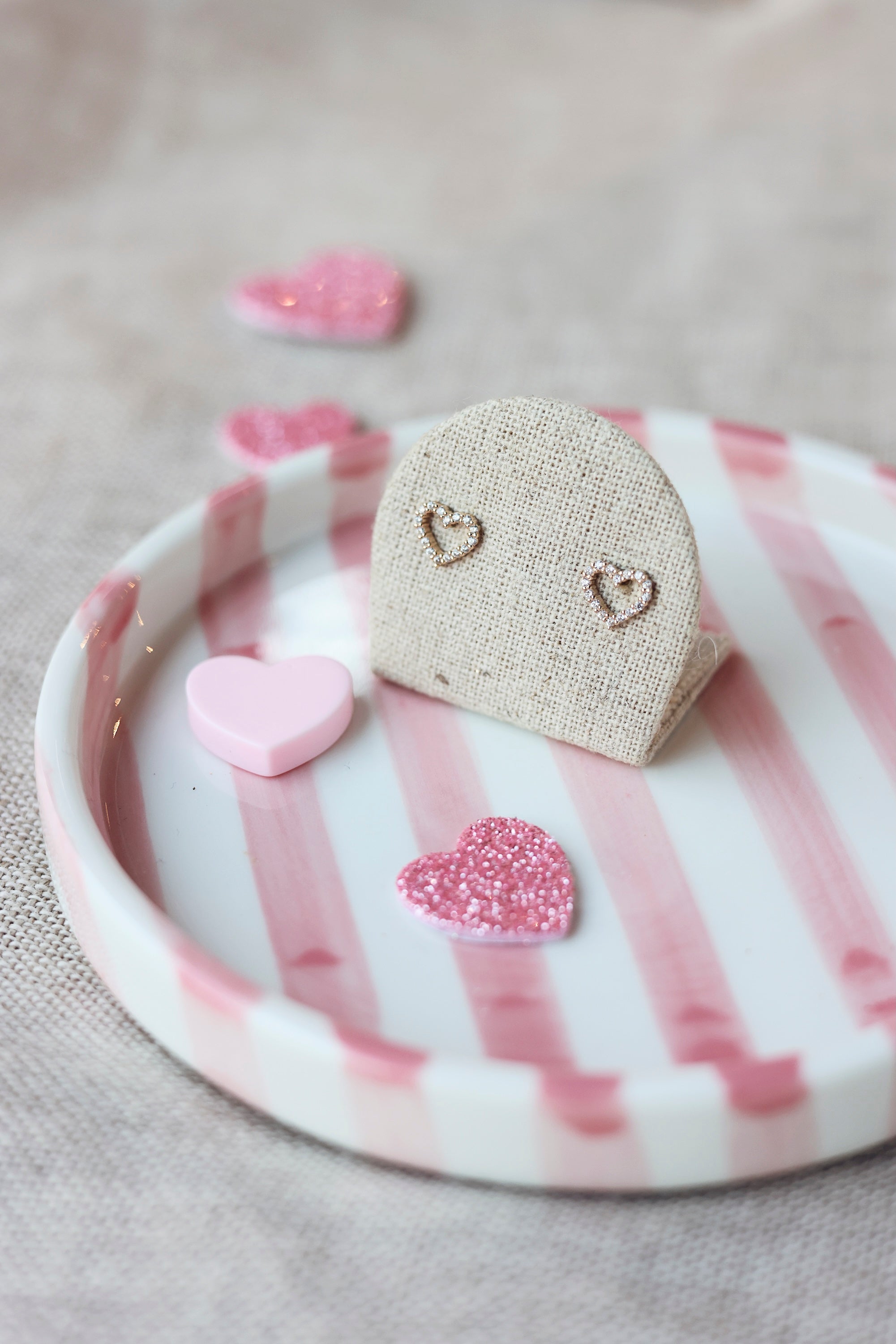 Boucles d'oreilles puces Alyssia dorées en forme de cœur ajouré ornées de zircons - Bijou photographié sur un présentoir en lin gris dans une ambiance saint valentin