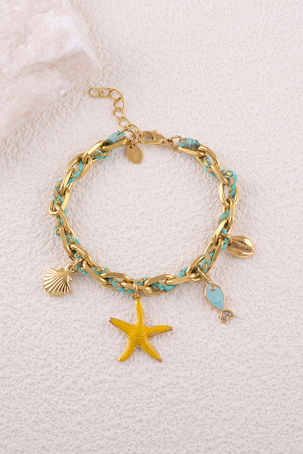 Bracelet doré Awen maille tressée avec cordon et pendentifs étoile de mer, coquillages et poisson - Bijou photographié sur fond en mur blanc