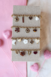 Bracelet doré Natty maille avec pendentifs coeurs, lune, fleur et trèfle - Bijou photographié sur un présentoir en lin gris