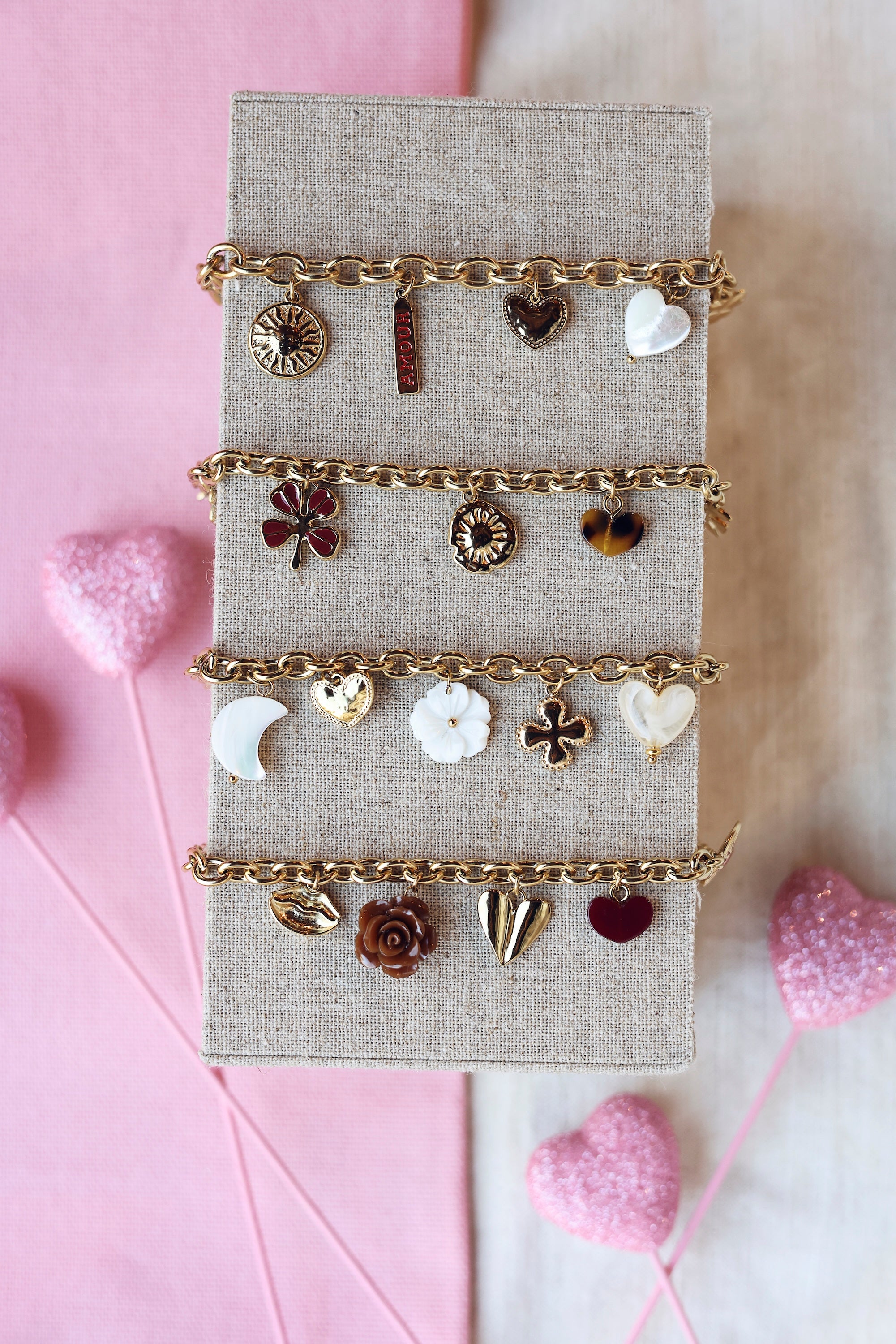 Bracelet doré Natty maille avec pendentifs coeurs, lune, fleur et trèfle - Bijou photographié sur un présentoir en lin gris