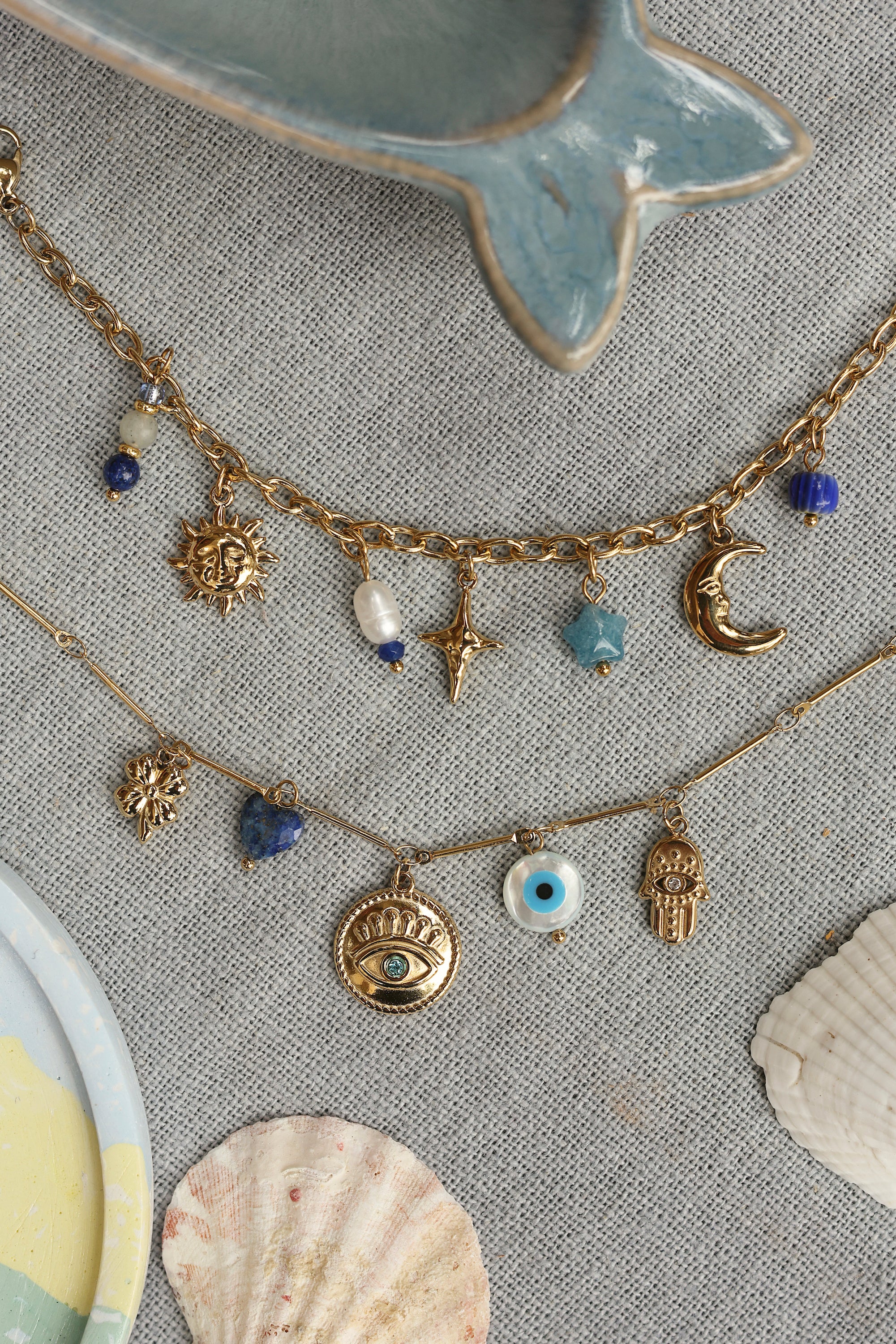 Bracelet doré Diena orné d’un cœur en sodalite, d’une médaille œil et d’une main chacune ornée d’un zircon, ainsi que d’un trèfle doré et d’un œil en nacre - Bijou photographié dans un décor estival 