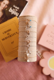 Bracelet personnalisable doré Lilou protect avec anneau torsadé et œil protecteur en nacre pour lettres de l'alphabet et charms - Bijou photographié sur un présentoir en lin avec d'autres bracelets personnalisables