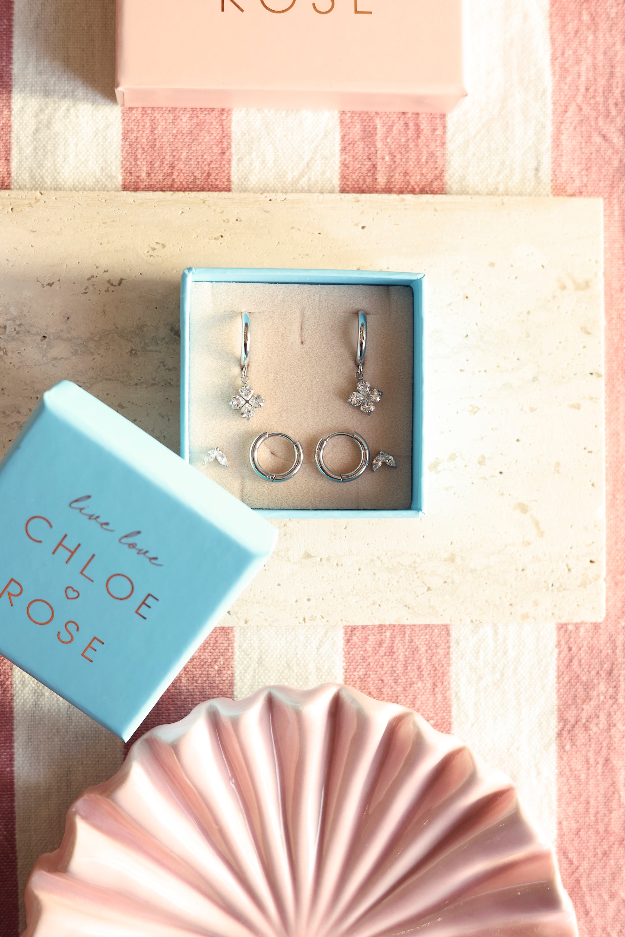 Coffret de parure de bijoux Flavia comprenant deux paires de mini créoles argentées et une paire de puces assorties - Bijoux photographiés dans un écrin à bijoux bleu posé sur une planche en marbre avec des décorations roses