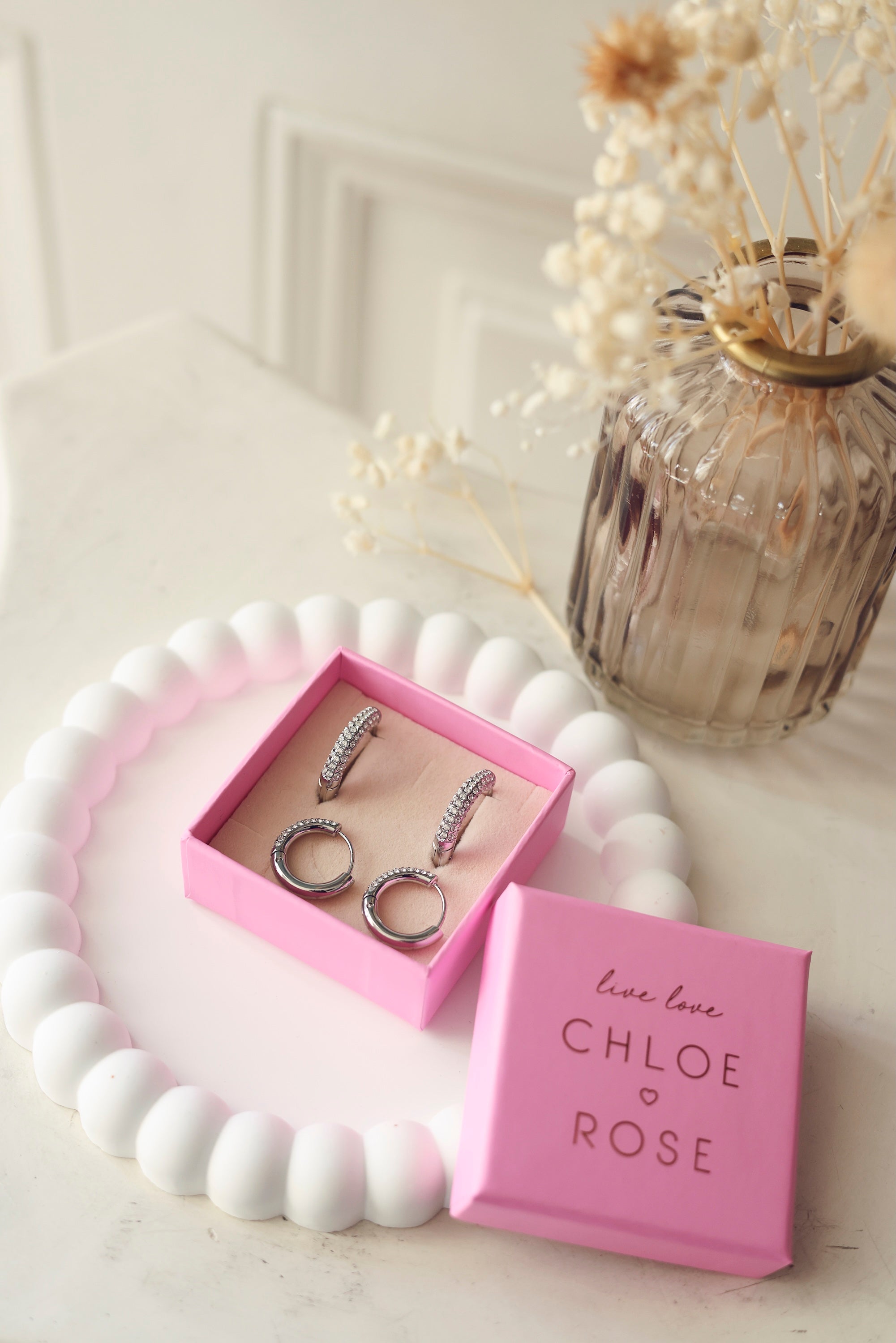 Coffret de parure de bijoux Lara contenant deux paires de créoles argentées ornées de zircons - Bijoux photographiés dans un écrin à bijoux rose posé dans un vide poche blanc avec des fleurs séchées