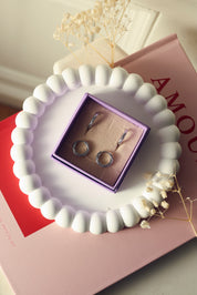 Coffret de parure de bijoux Laura contenant deux paires de mini créoles argentées torsadées - Bijoux photographiés dans un écrin à bijou lila dans un vide poche blanc posé sur un livre rose et rouge "amour" avec des fleurs séchées