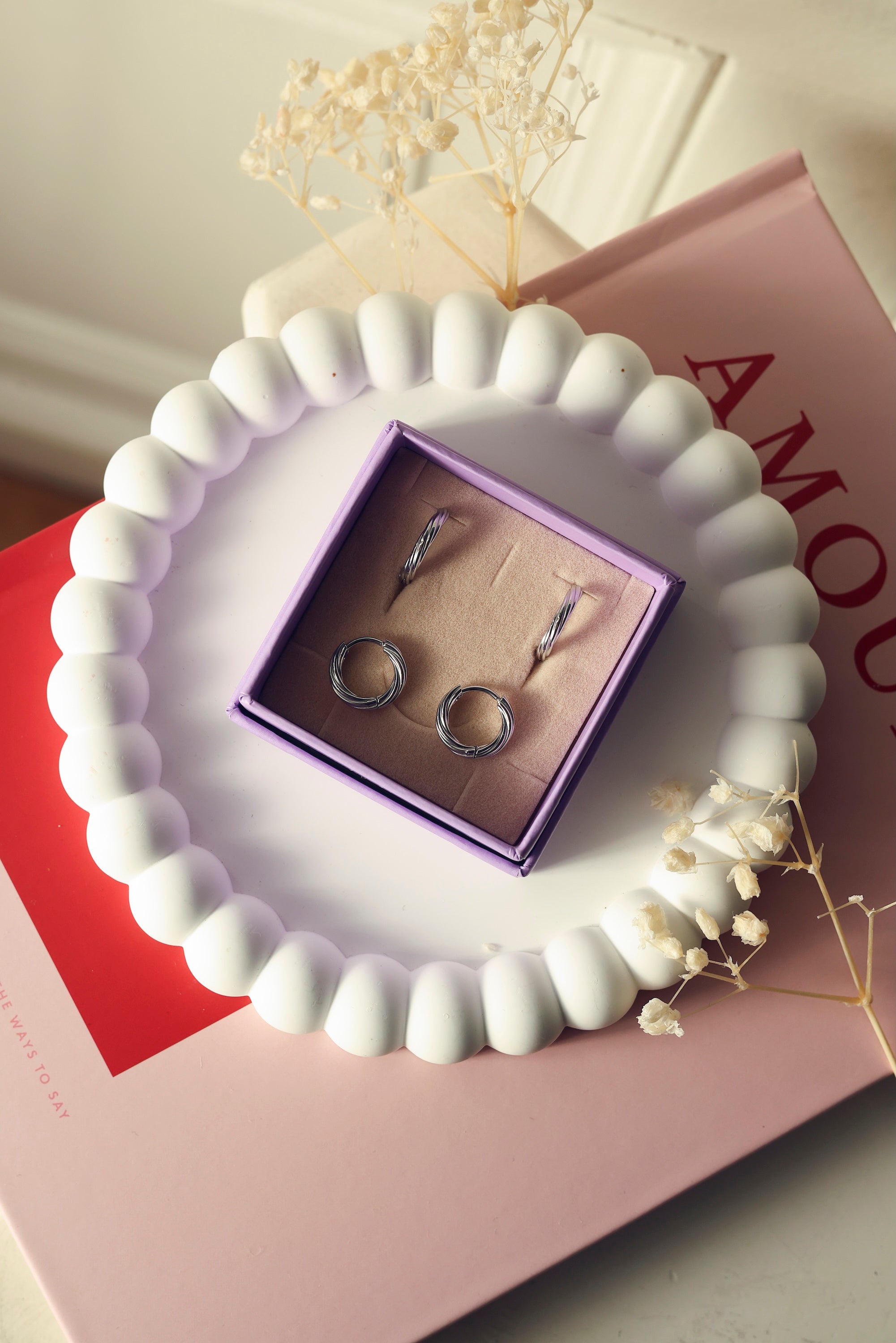 Coffret de parure de bijoux Laura contenant deux paires de mini créoles argentées torsadées - Bijoux photographiés dans un écrin à bijou lila dans un vide poche blanc posé sur un livre rose et rouge "amour" avec des fleurs séchées