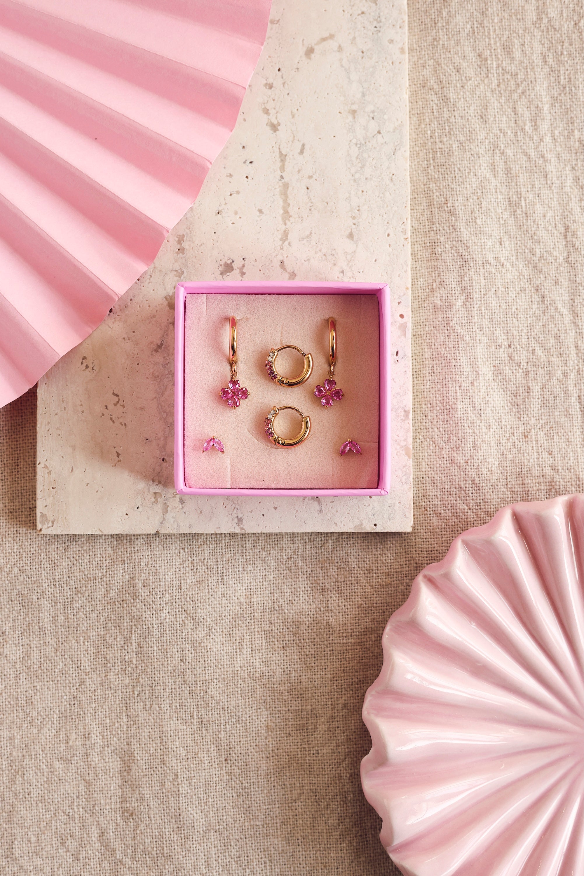 Coffret de parure de bijoux Sonia rose comprenant deux paires de mini créoles dorées ornées de zircons et une paire de puces assorties - Bijoux photographiés dans un écrin à bijou rose sur un support en marbre posé sur du lin avec des décoration rose
