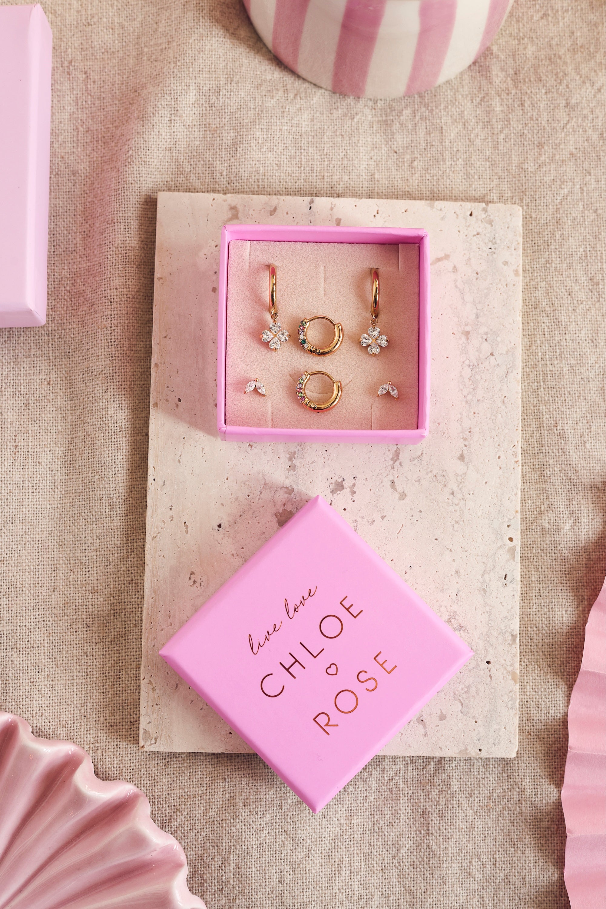 Coffret de parure de bijoux Sonia blanc comprenant deux paires de mini créoles dorées ornées de zircons et une paire de puces assorties - Bijoux photographiés dans un écrin à bijou rose sur un support en marbre posé sur du lin avec des décorations roses