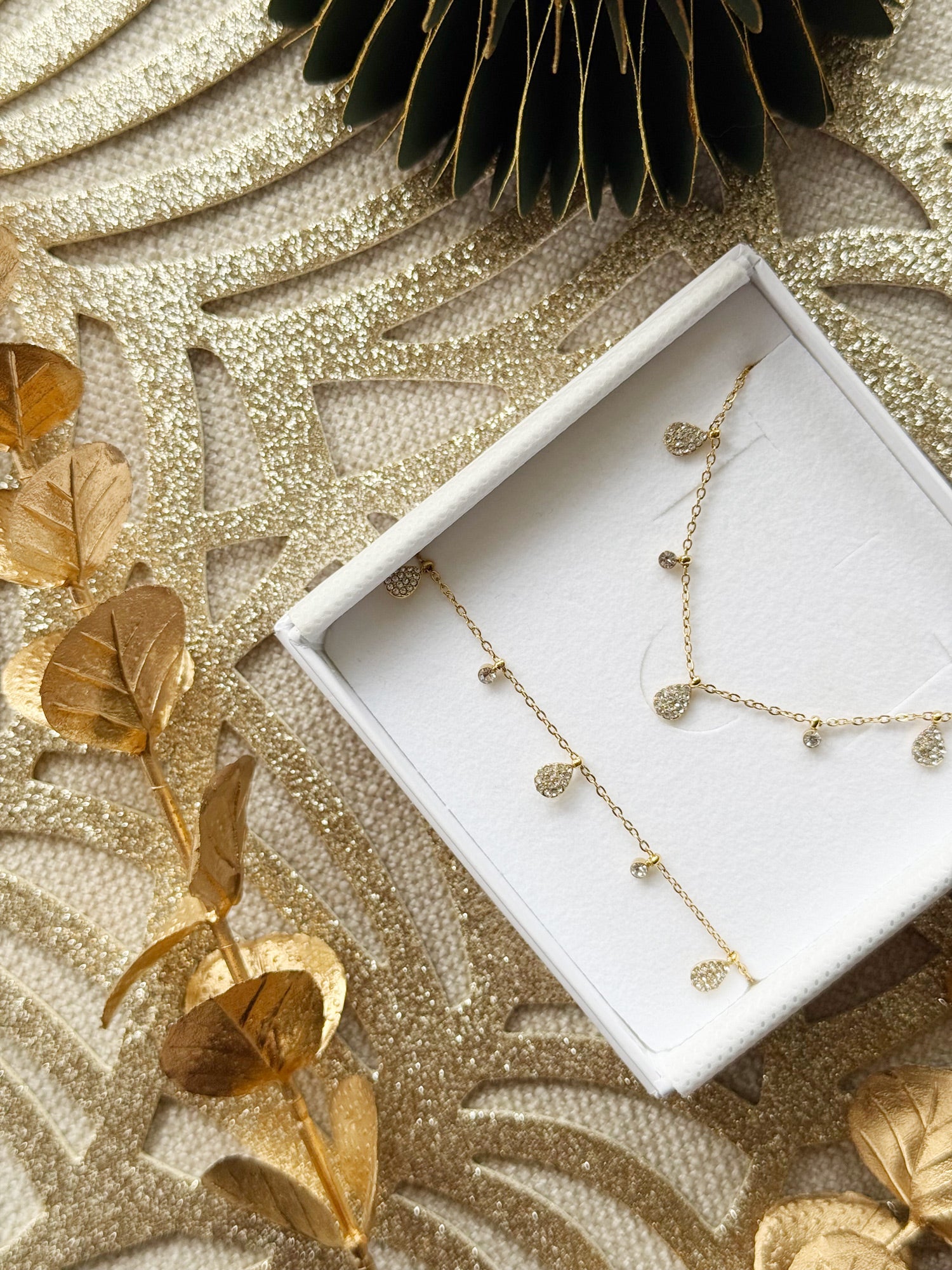Coffret de parure de bijoux Davia goutte contenant un collier et un bracelet dorés, ornés de petits charms en forme de goutte sertis de zircons - Bijoux photographiés dans un écrin à bijoux blanc dans une ambiance de Noël