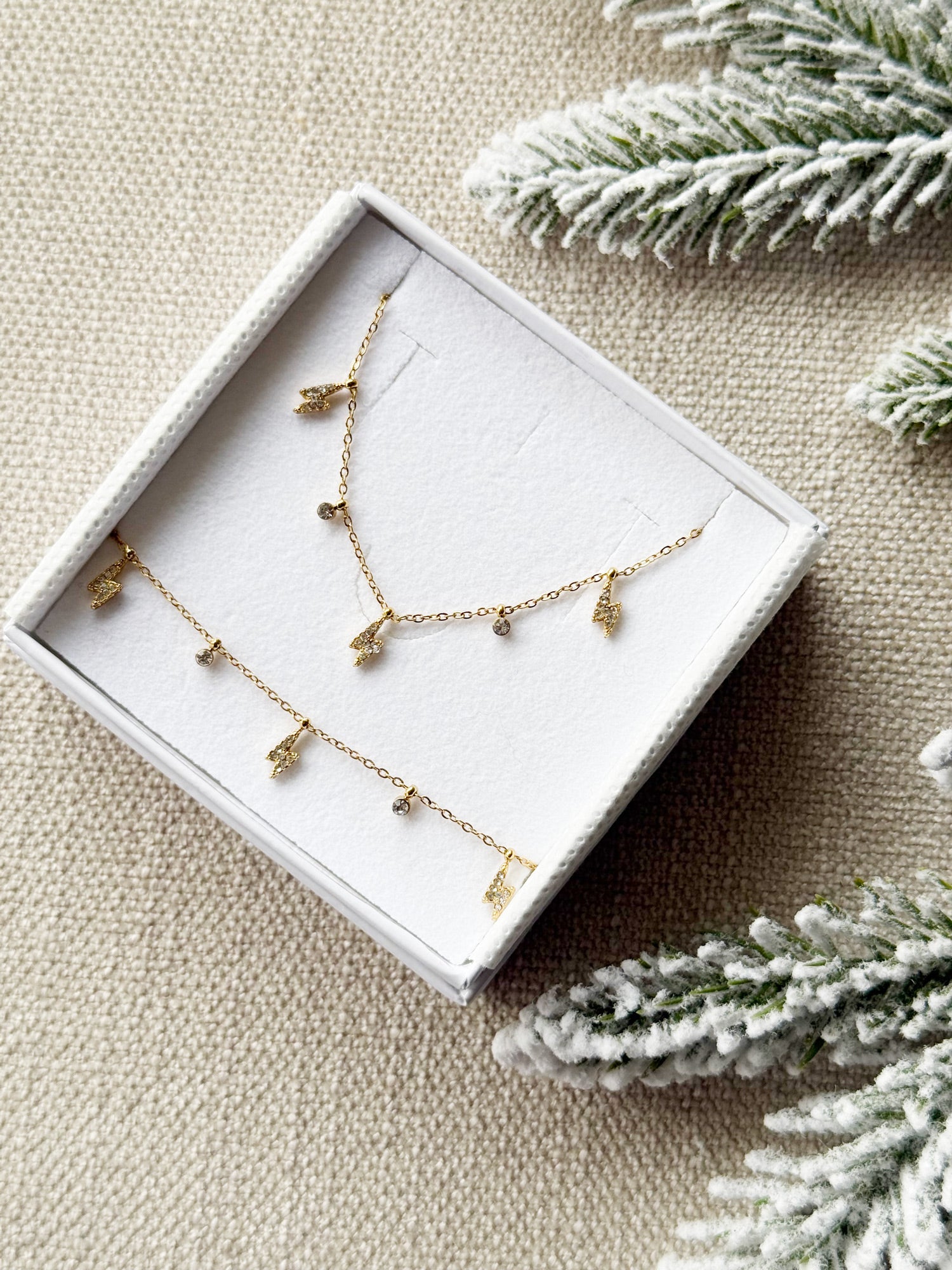 Coffret de parure de bijoux Davia éclair contenant un collier et un bracelet dorés, ornés de petits charms en forme d’éclair sertis de zircons - Bijoux photographiés dans un écrin à bijoux blanc dans une ambiance hivernale