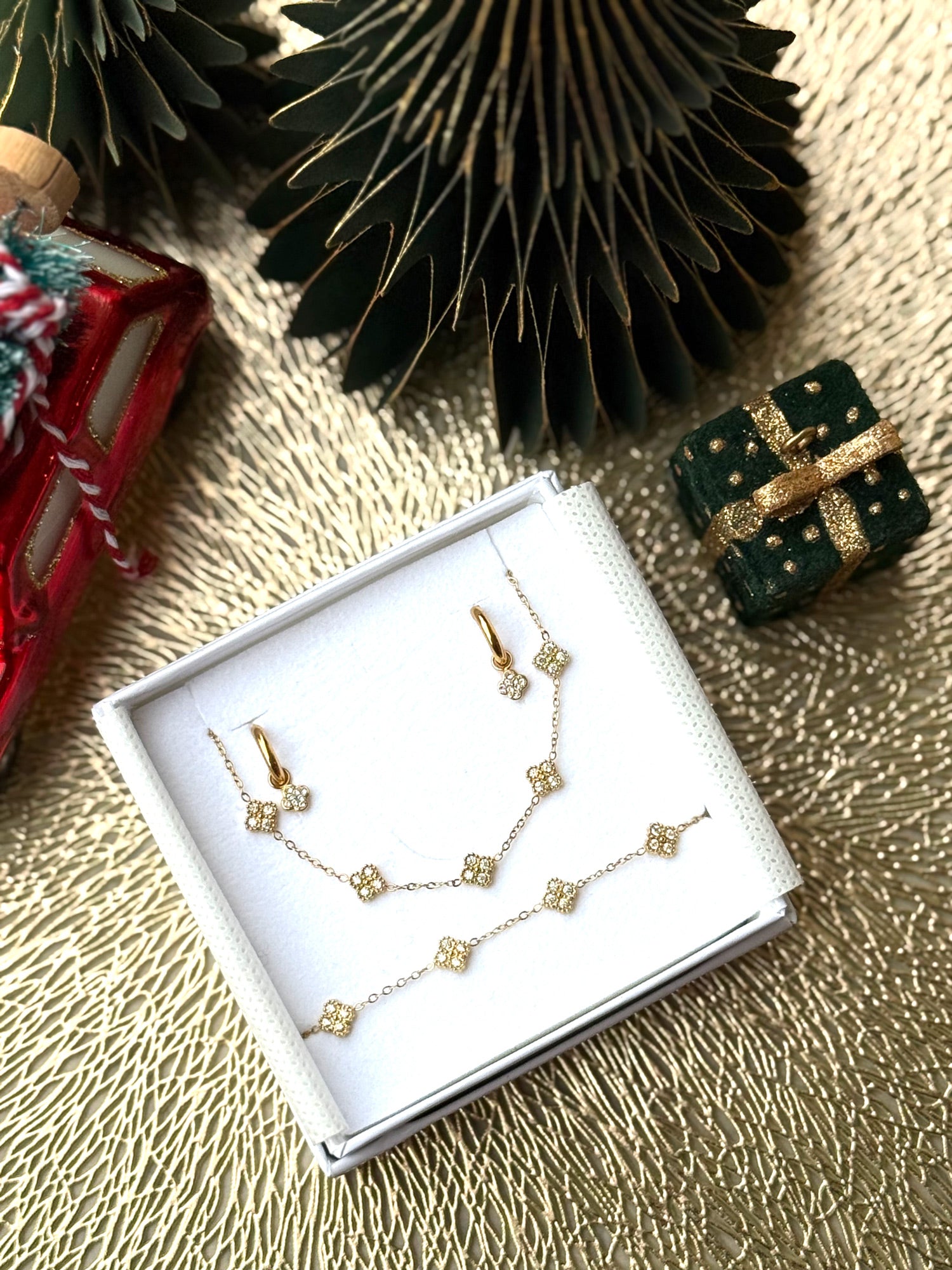 Coffret de parure Dolce comprenant une paire de mini créoles, un collier et un bracelet dorés, ornés de petites fleurs serties de zircons - Bijoux photographiés dans un écrin à bijoux blanc dans une ambiance de Noël
