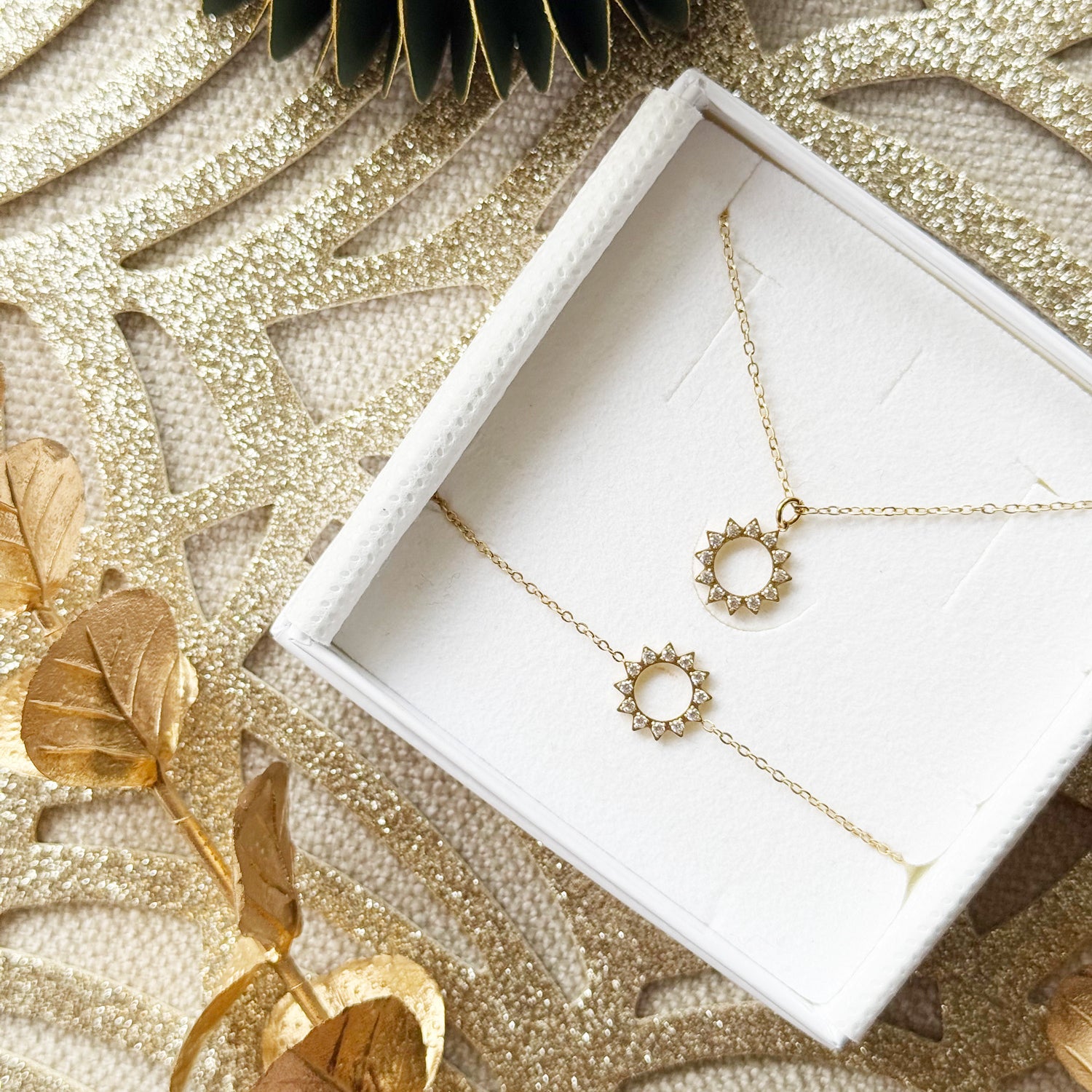 Coffret de parure de bijoux Joy contenant un collier et un bracelet dorés, ornés d’un soleil serti de zircons - Bijoux photographiés dans un écrin à bijoux blanc dans une ambiance de Noël