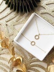 Coffret de parure de bijoux Joy contenant un collier et un bracelet dorés, ornés d’un soleil serti de zircons - Bijoux photographiés dans un écrin à bijoux blanc dans une ambiance de Noël