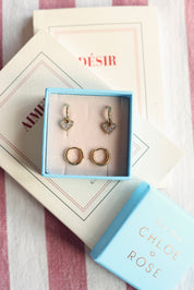 Coffret de parure de bijoux Lily contenant deux paires de mini créoles dorées, une paire simple et une paire avec pendentif cœur serti de zircons - Bijoux photographiés dans un écrin à bijoux bleu posé sur des livres