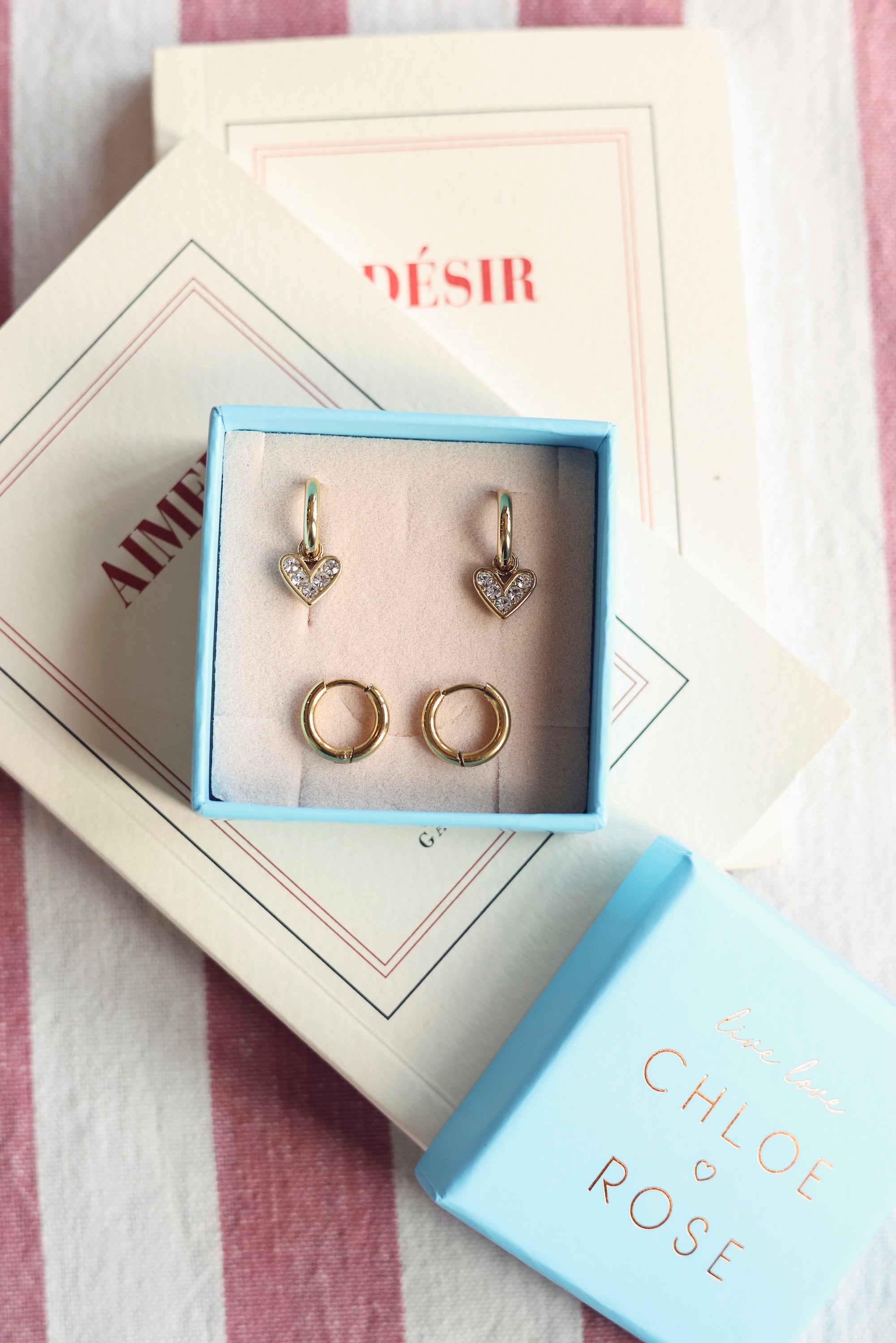 Coffret de parure de bijoux Lily contenant deux paires de mini créoles dorées, une paire simple et une paire avec pendentif cœur serti de zircons - Bijoux photographiés dans un écrin à bijoux bleu posé sur des livres