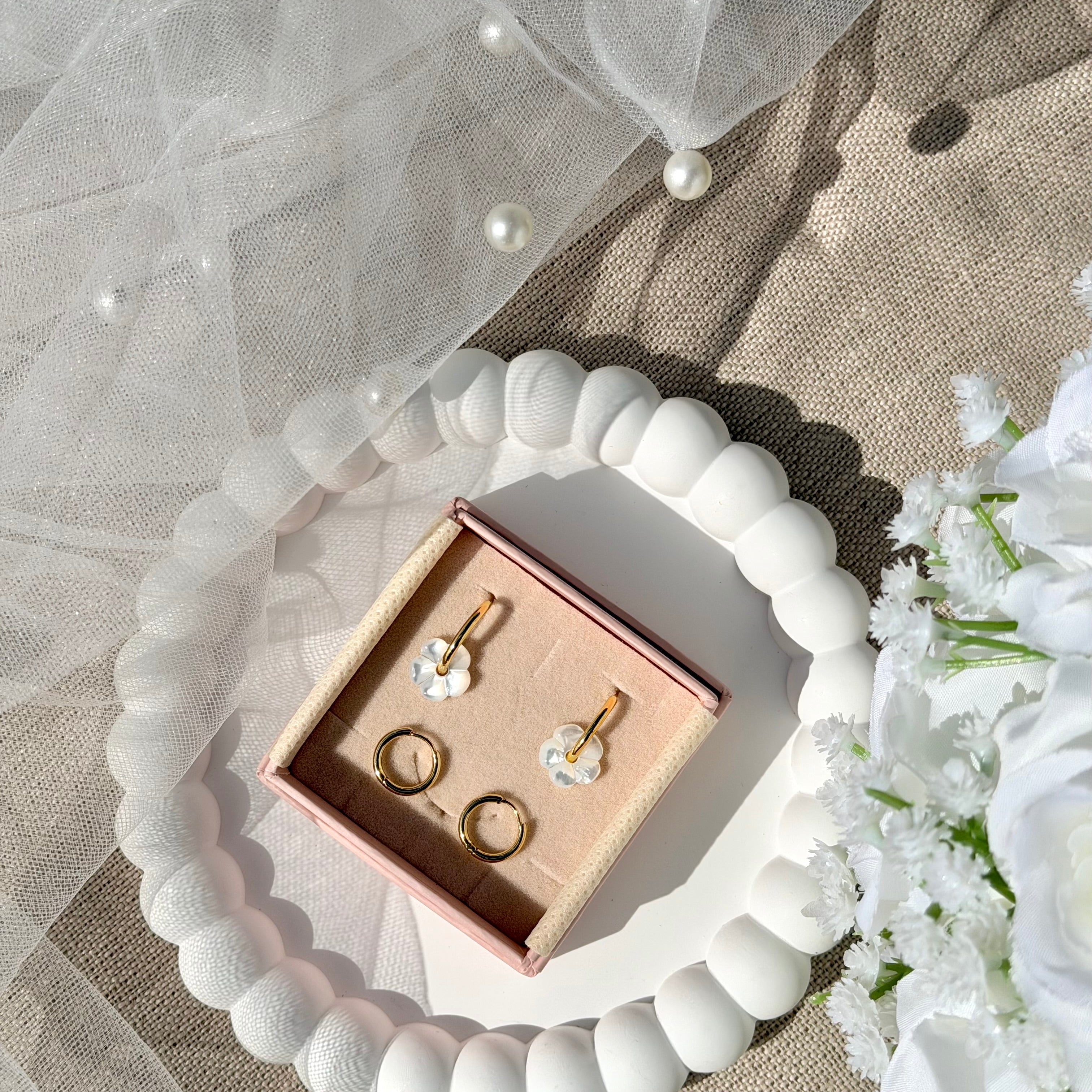 Coffret de parure de bijoux Lucille comprenant une paire de mini créoles dorées ornées d’une fleur en nacre ainsi qu’une paire de créoles simples - Coffret photographié dans une coupole blanche avec un voile de mariée et un bouquet