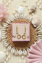 Coffret de parure de bijoux Oasis blanc comprenant une paire de mini créoles dorées ornées d’un pendentif pétales serti de zircons ainsi qu’une paire de créoles simples et une paire de puces assorties - Bijoux photographiés dans un décor estival