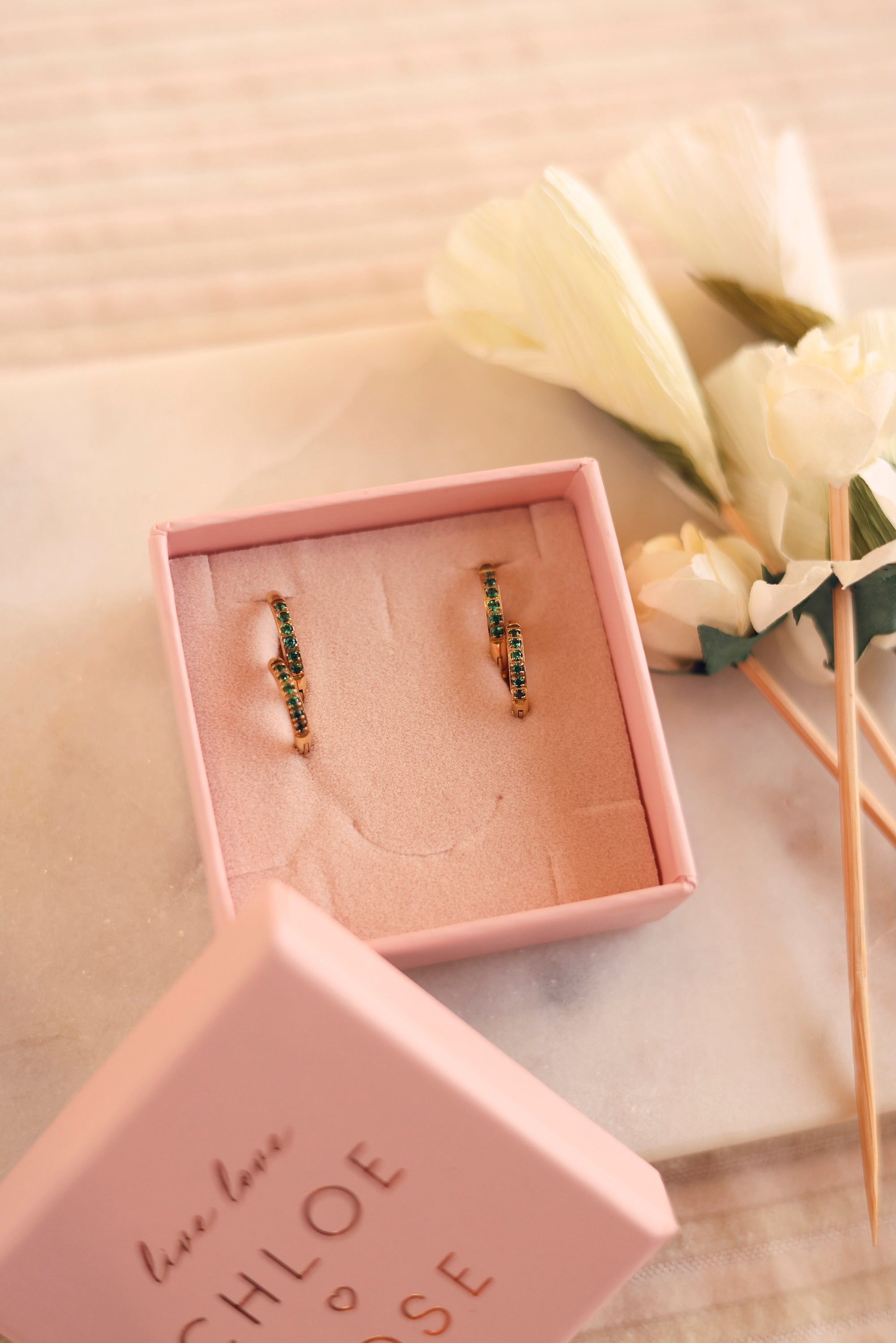 Coffret de parure de bijoux Sara vert contenant deux paires de mini créoles dorées ornées de zircons - Bijoux photographiés dans un écrin à bijoux rose posé sur du marbre blanc avec des fleurs