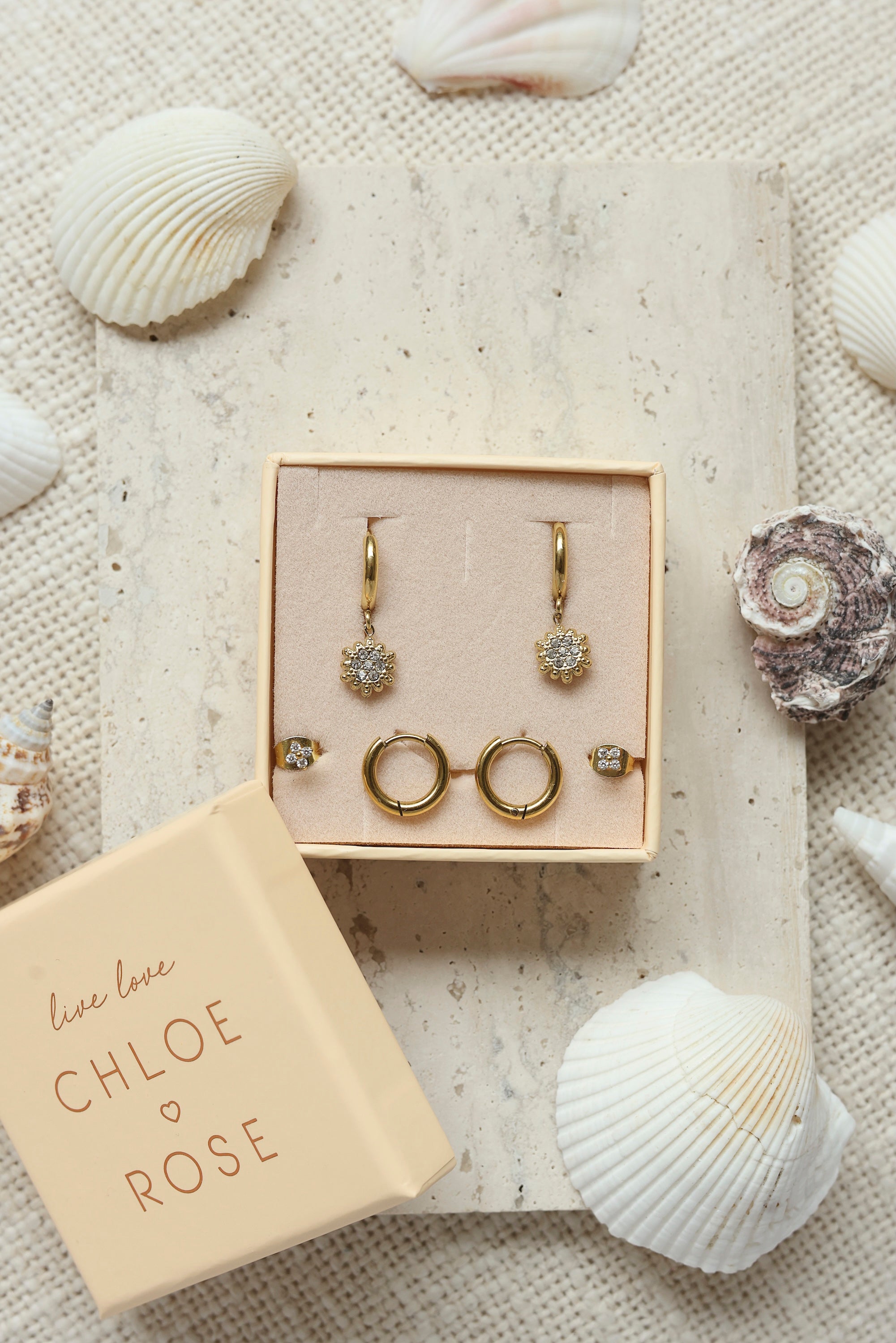Coffret de parure de bijoux Sunny comprenant une paire de mini créoles dorées ornées d’un soleil serti de zircons ainsi qu’une paire de créoles simples et une paire de puces en forme de fleur - Bijoux photographiés dans un décor estival
