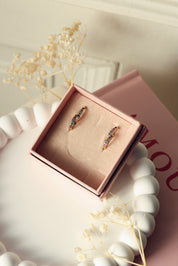 Coffret de parure de bijoux Sara multicolore contenant deux paires de mini créoles dorées ornées de zircons - Bijoux photographiés dans un écrin à bijou rose posé dans un vide poche blanc avec des fleurs séchées