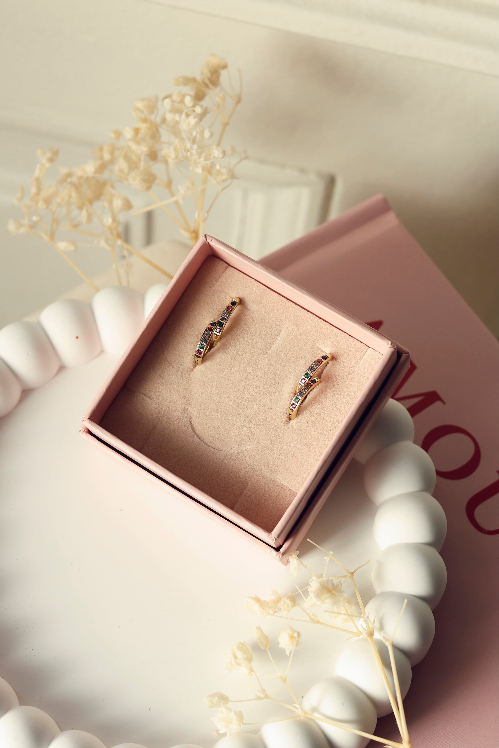 Coffret de parure de bijoux Sara multicolore contenant deux paires de mini créoles dorées ornées de zircons - Bijoux photographiés dans un écrin à bijou rose posé dans un vide poche blanc avec des fleurs séchées