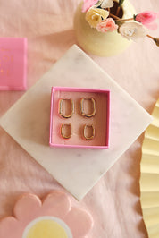Coffret de parure de bijoux Sophie contenant deux paires de mini créoles dorées rectangulaires aux bords arrondis - Bijoux photographiés dans un écrin à bijoux rose sur un bloc en marbre blanc avec des décorations roses et jaunes